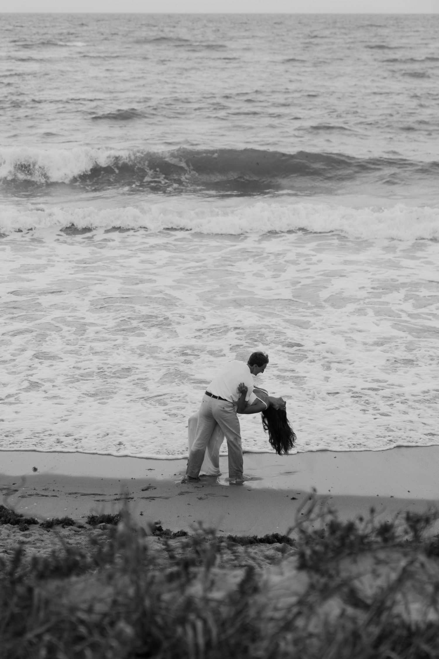 Black and white photo of a couple dancing romantically on a beach with ocean waves crashing behind them.