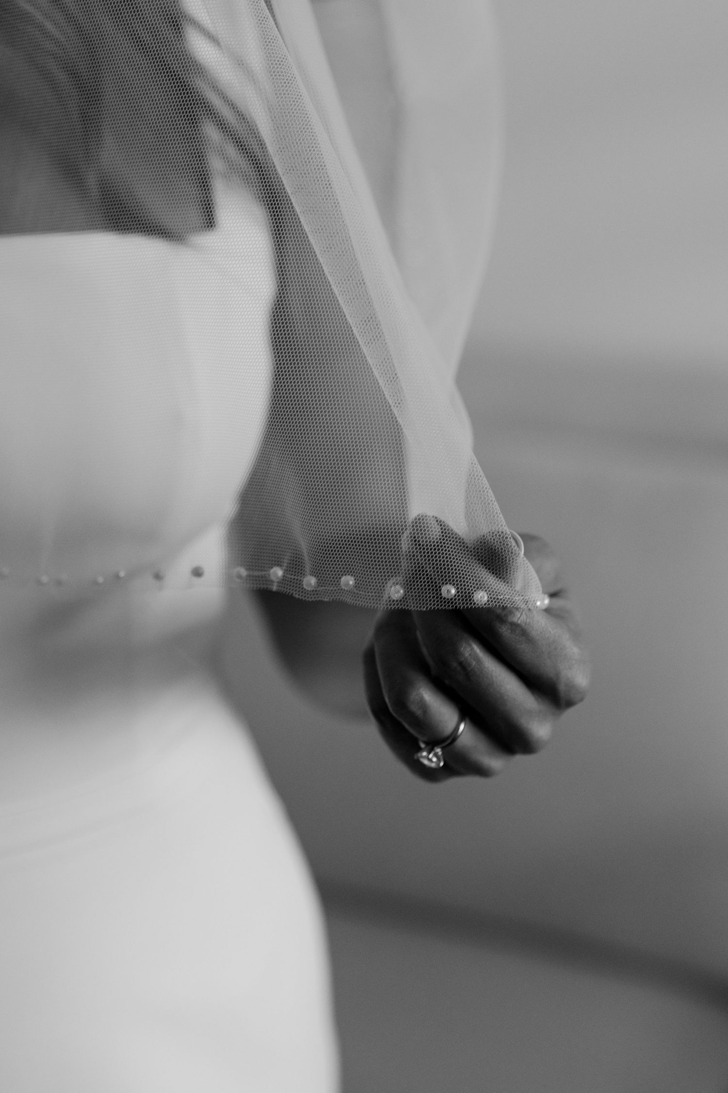 Black and white close-up of a bride holding her pearl-trimmed veil with an engagement ring visible.