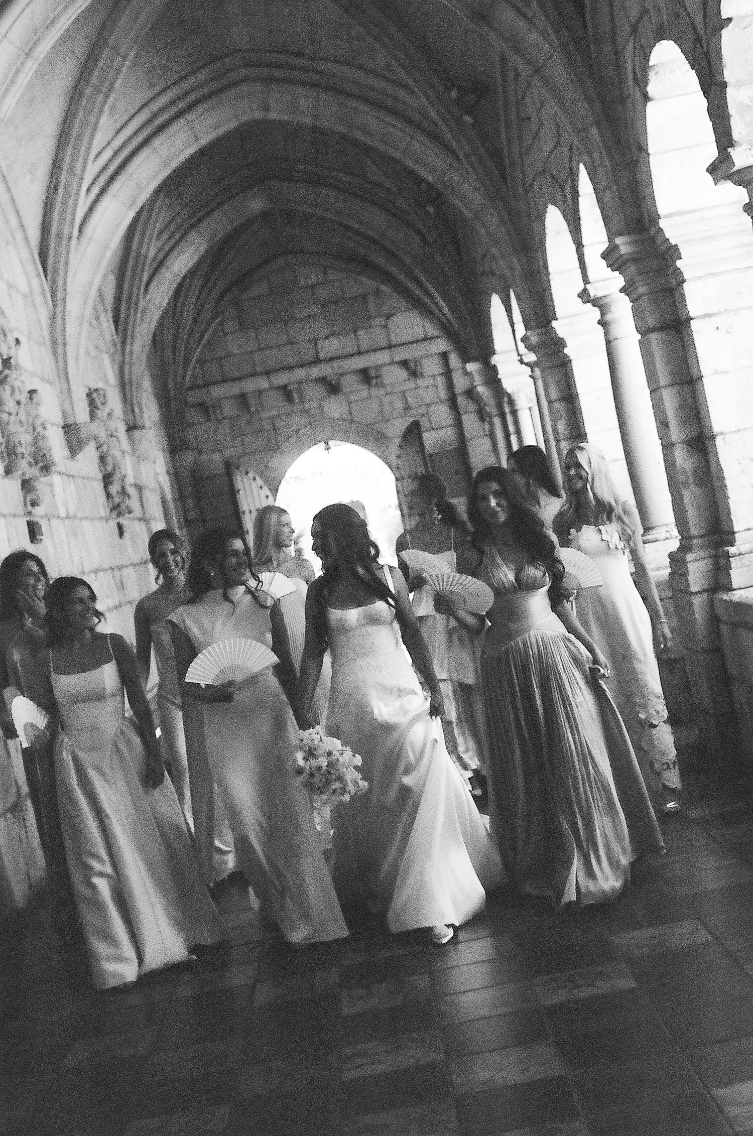 Black and white photo of a bride and bridesmaids walking through a stone archway corridor in elegant gowns.