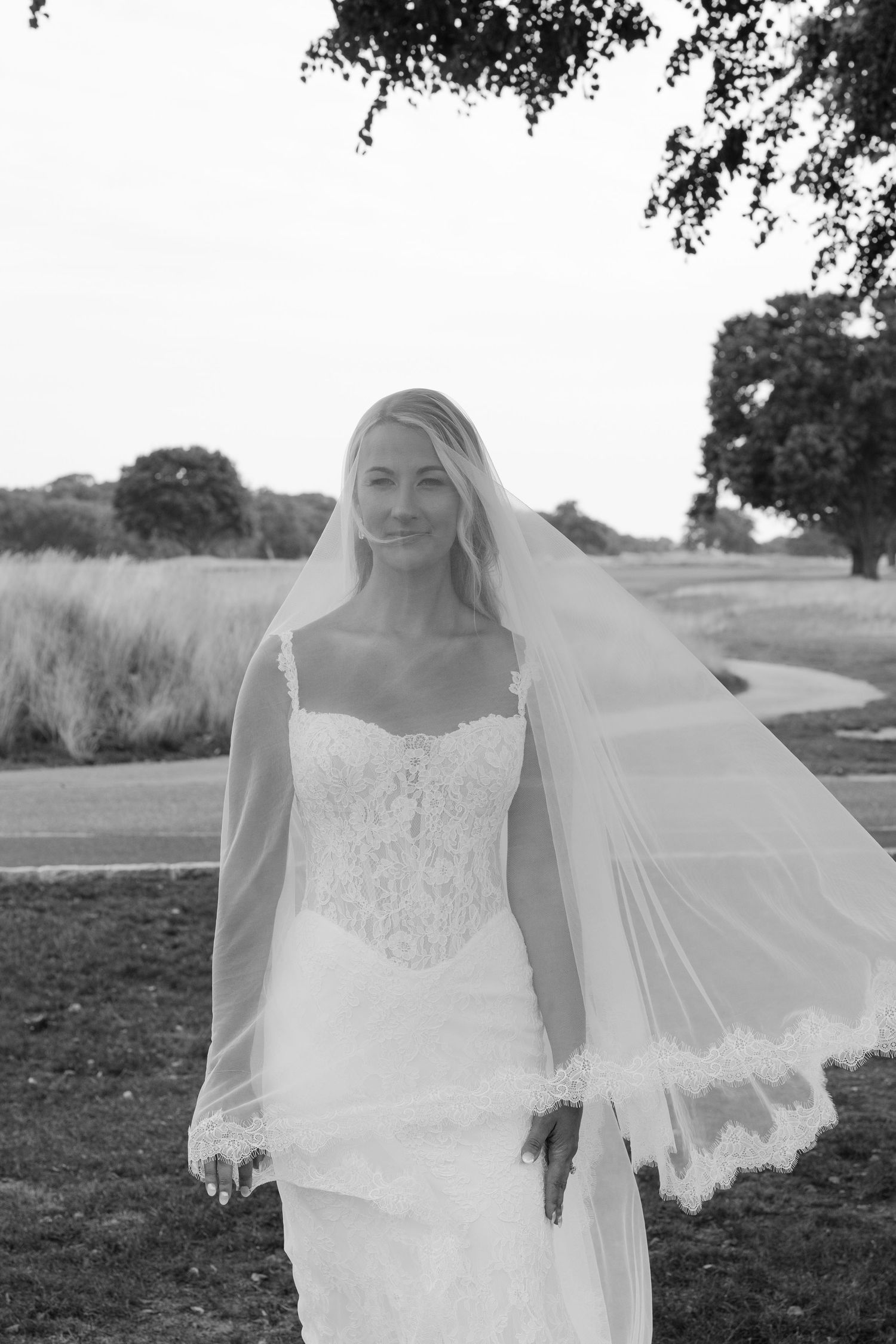 Elegant blonde bride in lace wedding gown and flowing veil poses outdoors in black and white portrait.