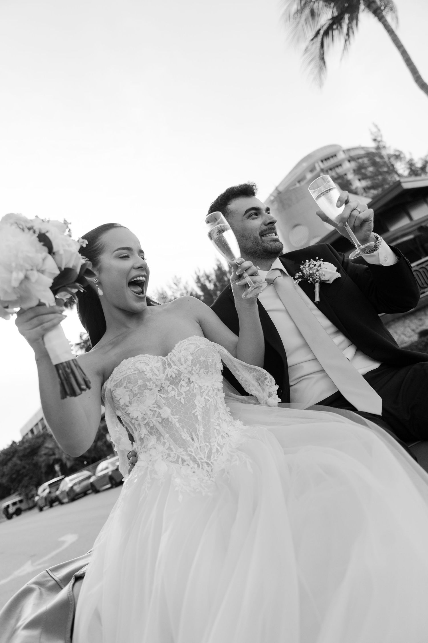 Joyful bride and groom celebrating outdoors with champagne glasses, bride holding bouquet in stunning lace ballgown.
