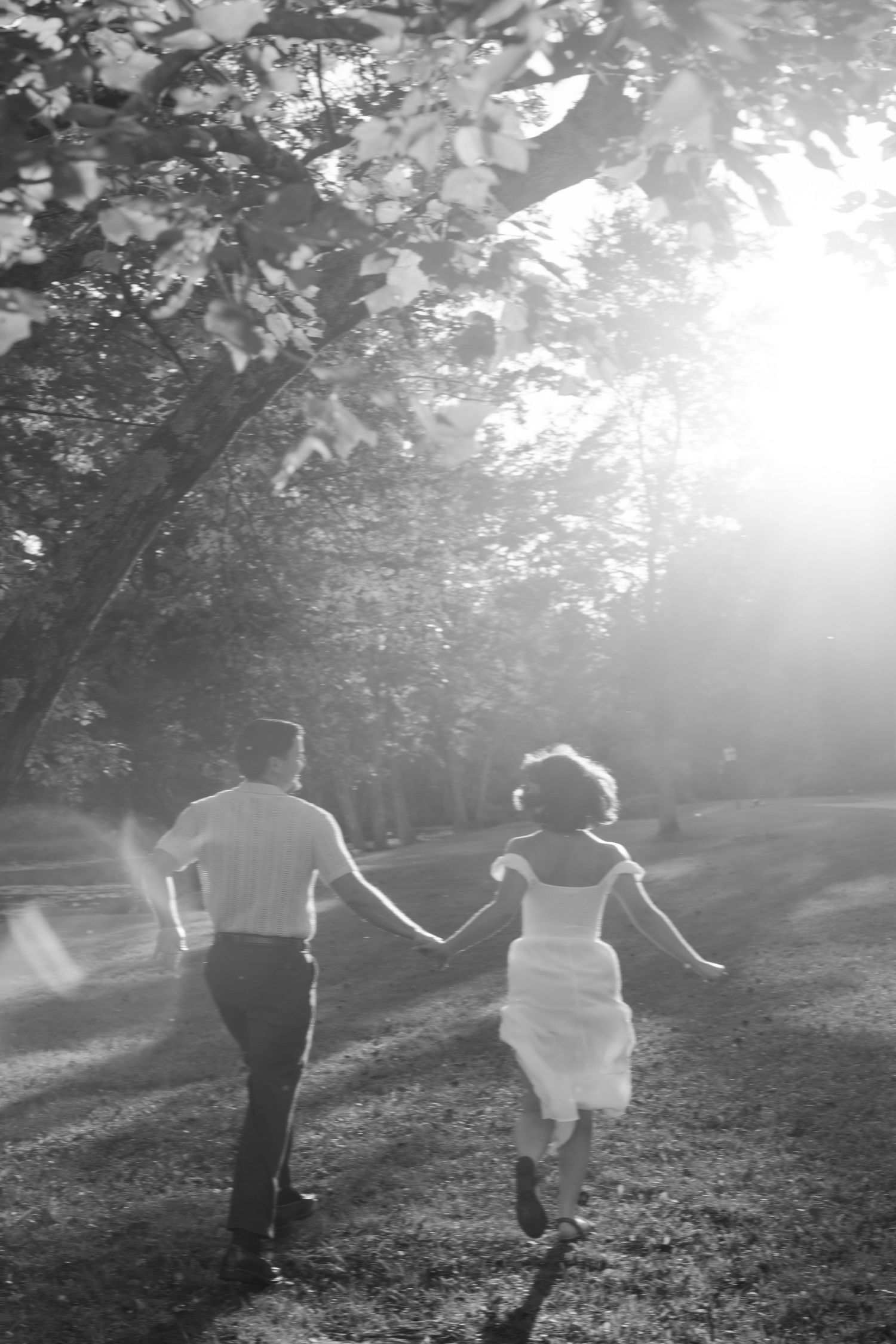 Black and white photo of a bride and groom holding hands, running through a sunlit park on their wedding day.