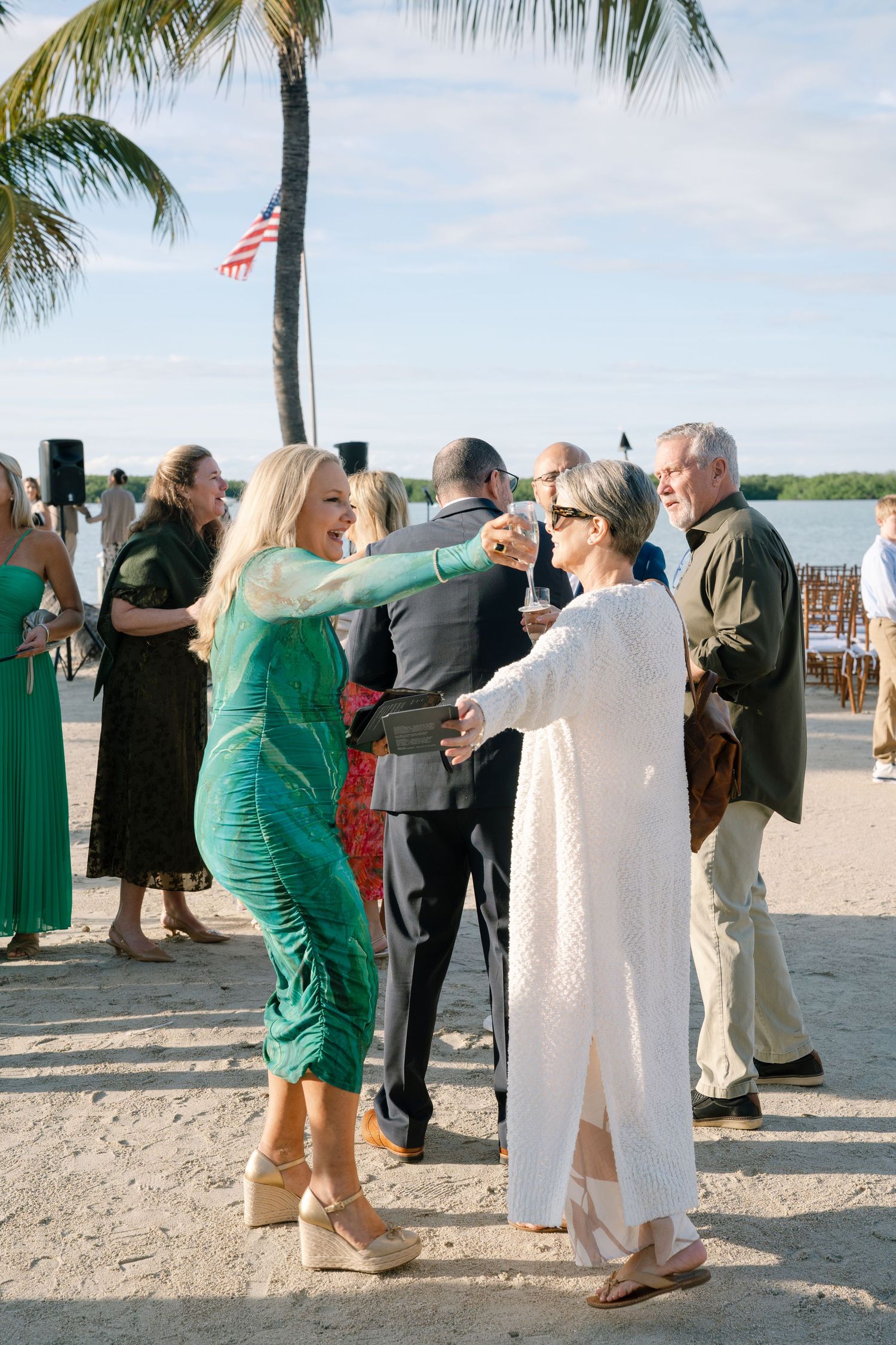 Wedding guests socialize on a beach with palm trees and an American flag.