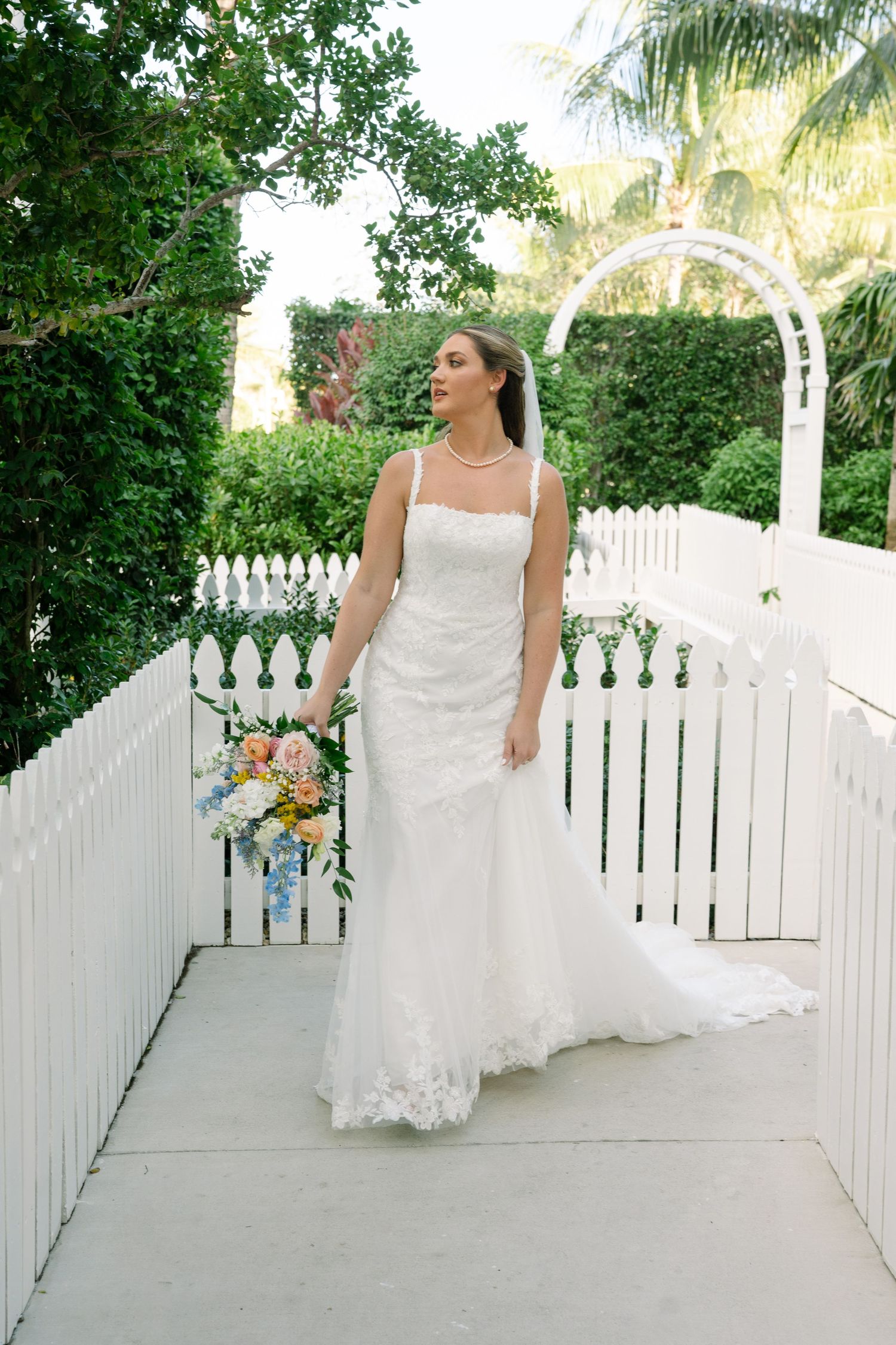 A bride in a white lace dress poses along a white picket fence walkway with tropical palm trees in the background.