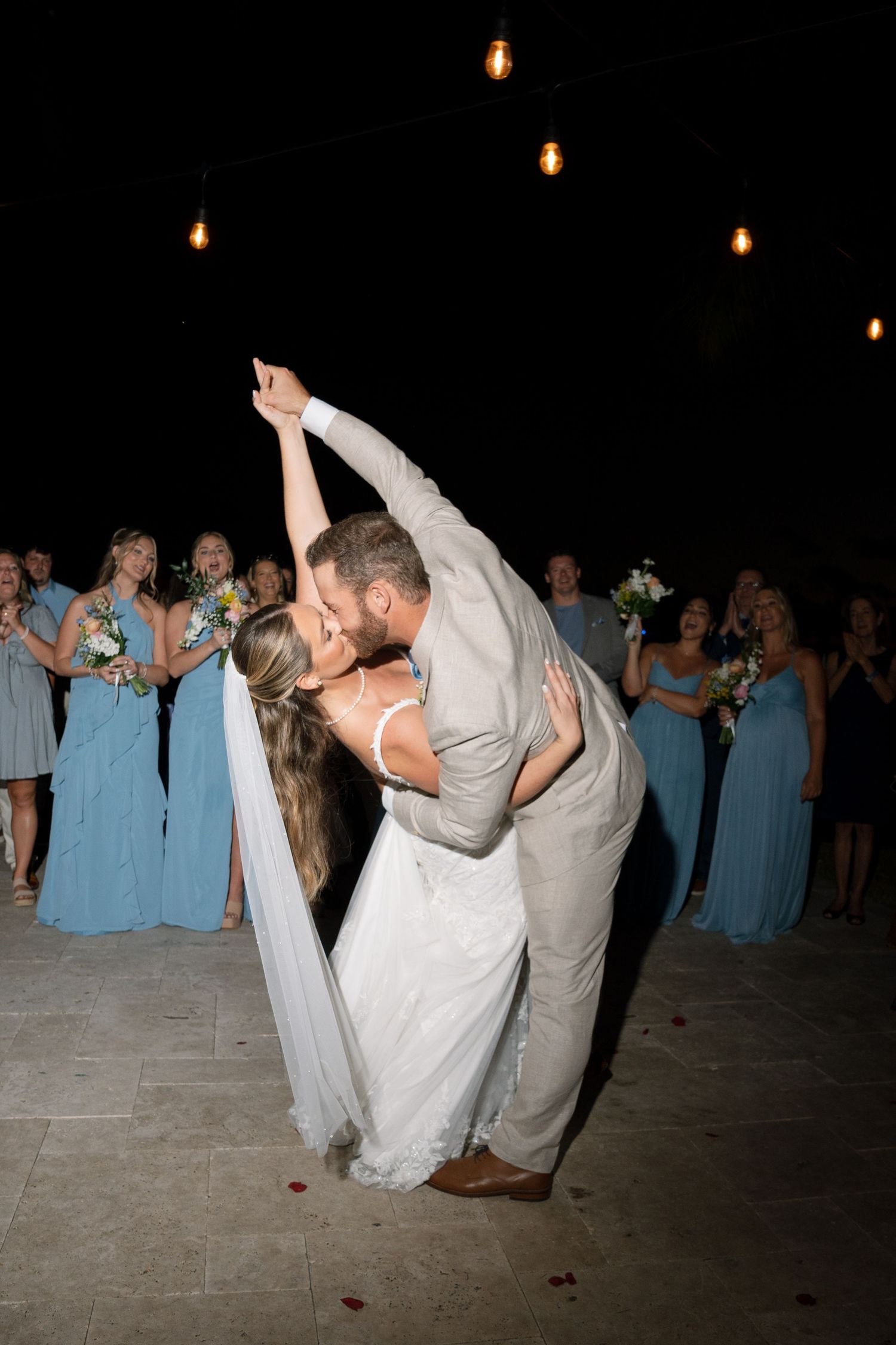 A romantic wedding dance dip on a dark dance floor surrounded by guests in light blue bridesmaid dresses.
