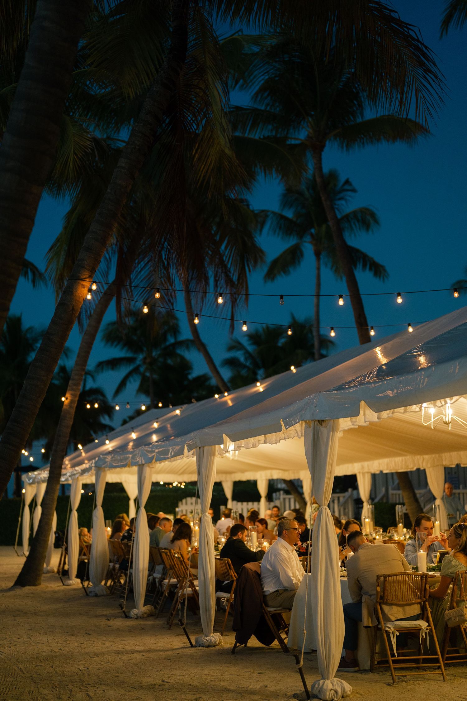 White tented outdoor beach reception at dusk with string lights and palm trees creating a romantic tropical atmosphere.