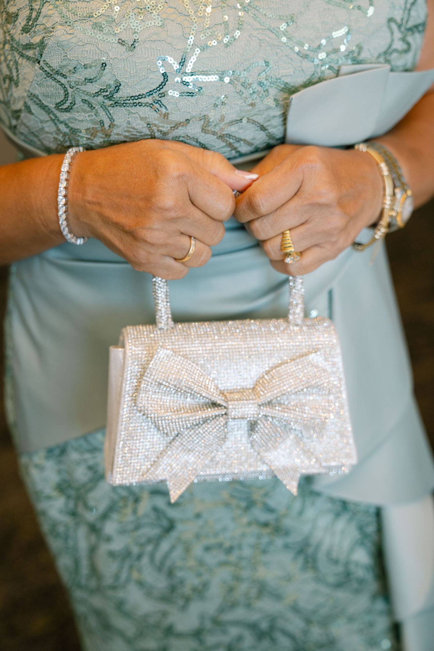 Hands holding a sparkly silver evening clutch with bow detail and pearl bracelet accessories.