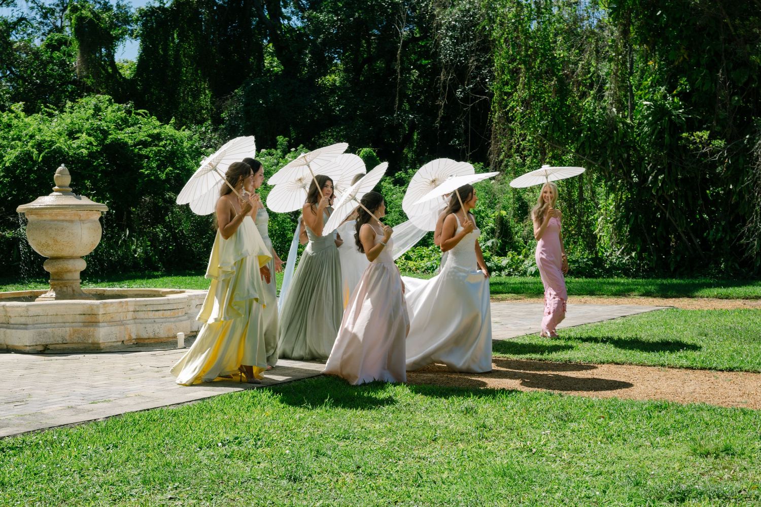 Bridesmaids in colorful gowns holding white parasols walk through a lush garden near a stone fountain.