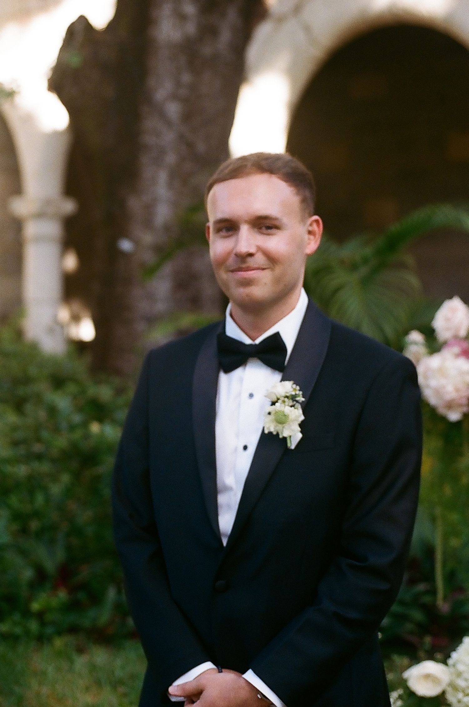 Smiling groom in classic black tuxedo with white boutonniere standing in a lush garden courtyard.