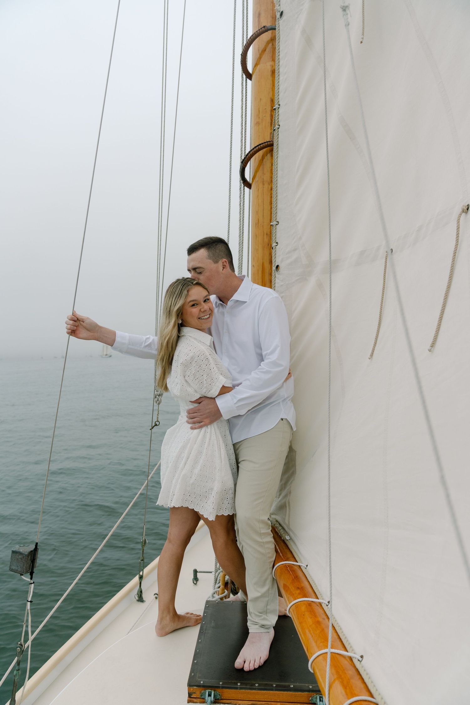 Couple embracing on a sailboat deck, woman in white dress and man in white shirt, with calm ocean in the background.