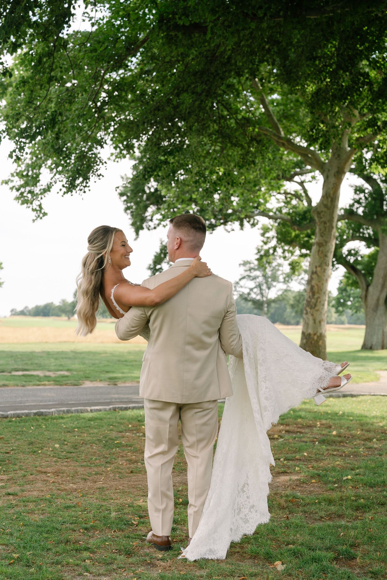 Groom lifts his smiling bride in a lace wedding gown under green trees on a sunny outdoor wedding day.