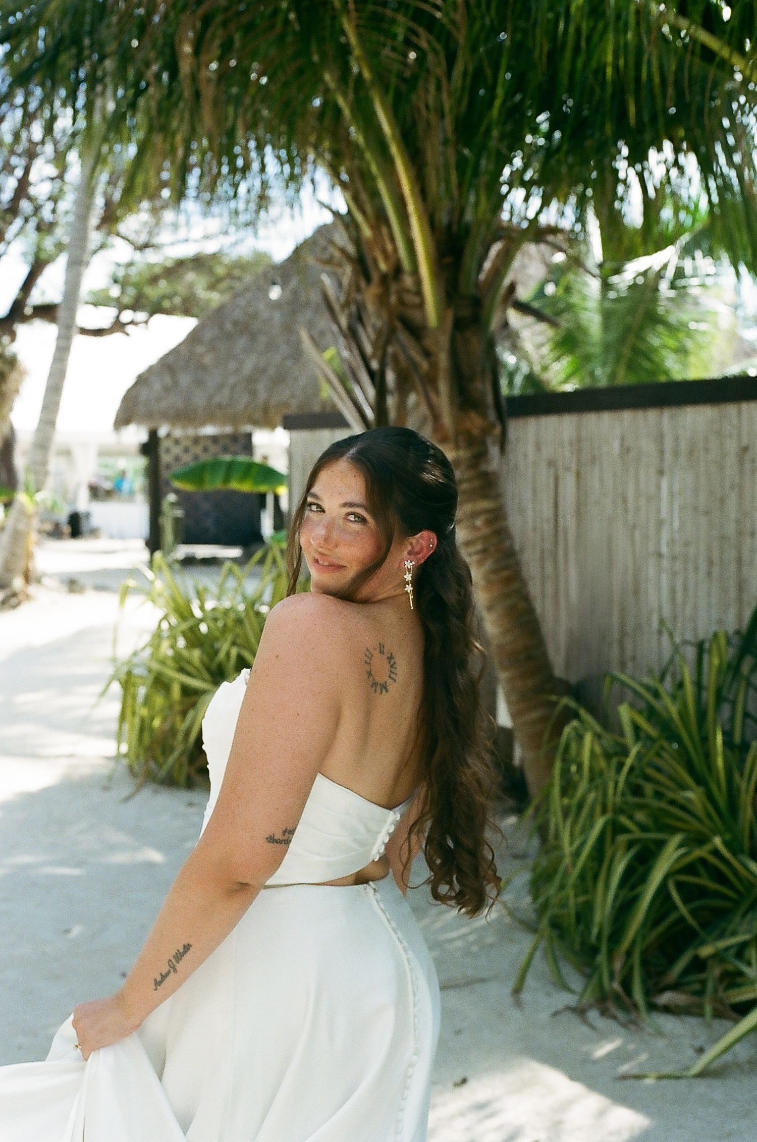 A woman in a white dress stands in front of a tropical setting with palm trees.