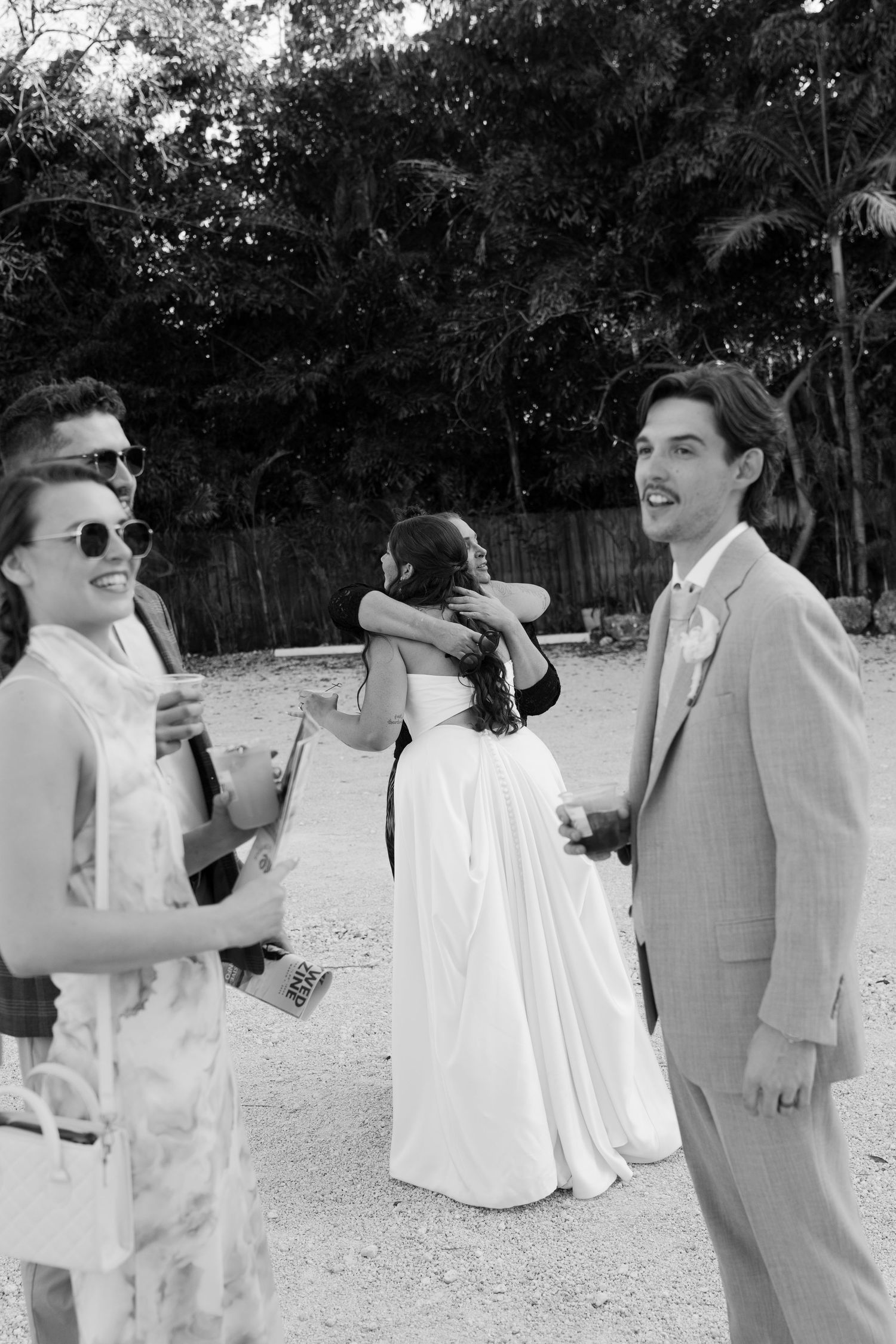 A black and white image shows a couple in formal attire walking together on a path in a garden setting.