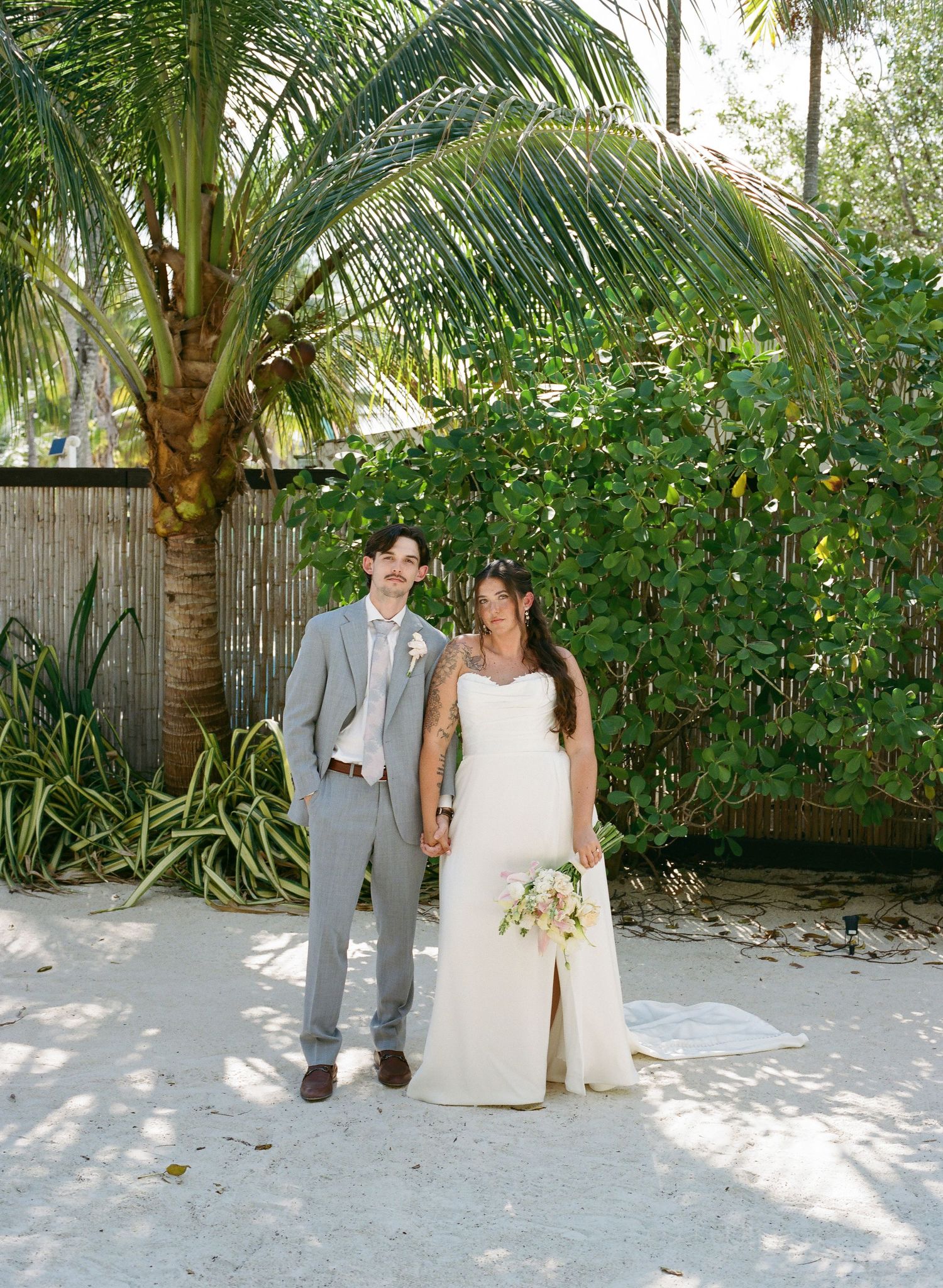 Bride and groom pose hand-in-hand on sandy beach surrounded by lush tropical palm trees and greenery.