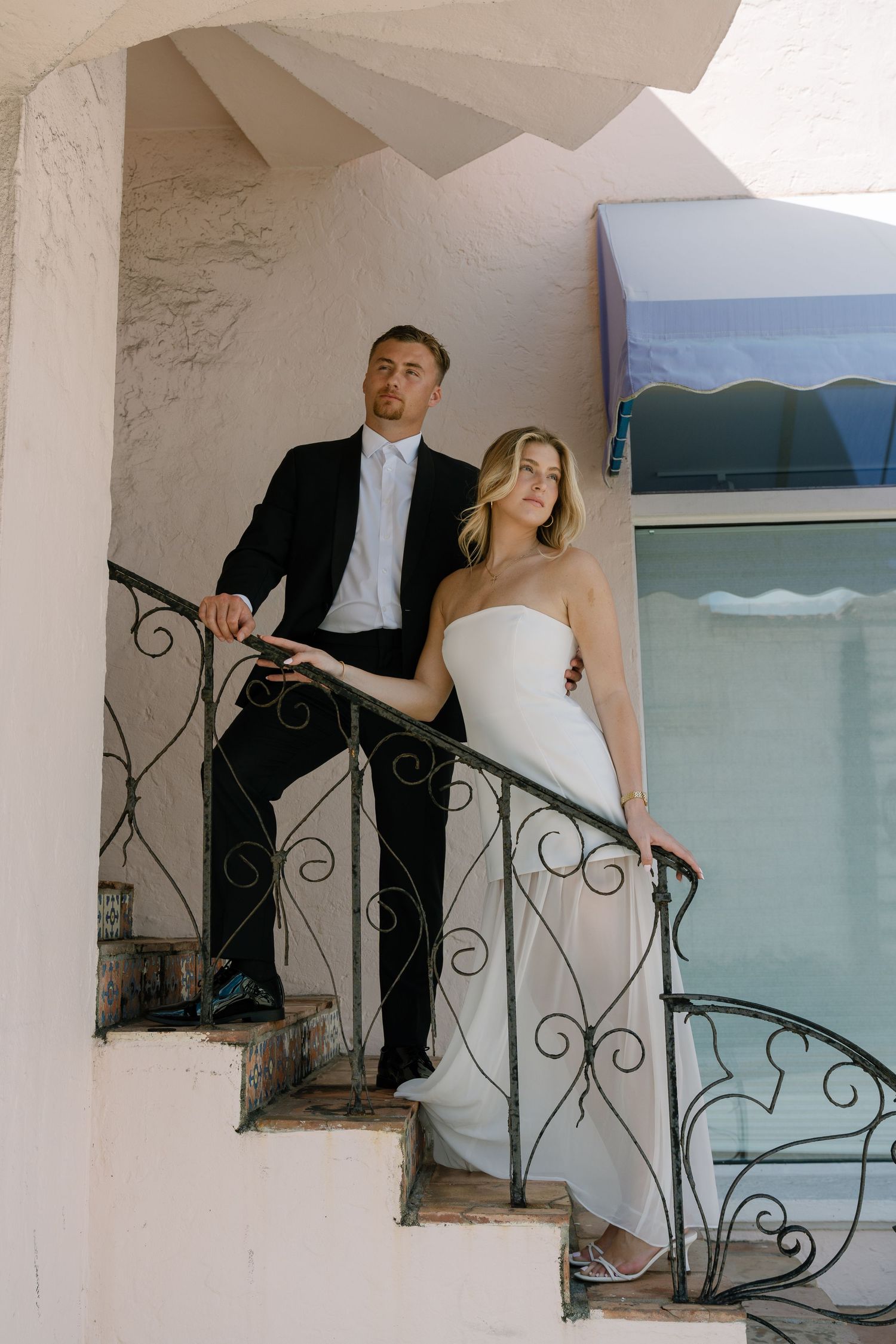 Bride and groom pose on ornate wrought iron staircase, she in strapless white gown, he in black suit.