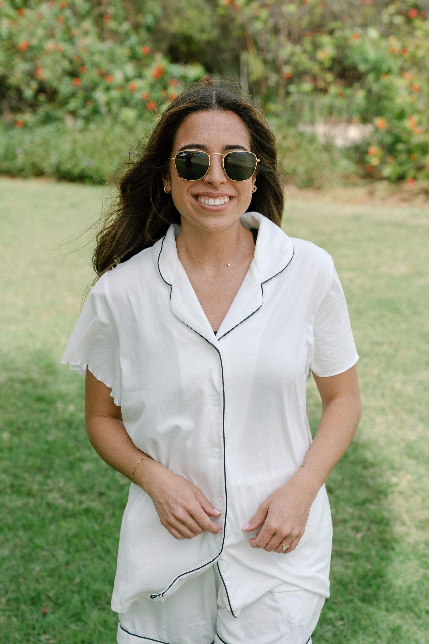 Smiling bride in white pajama shirt and sunglasses standing in a lush green garden.