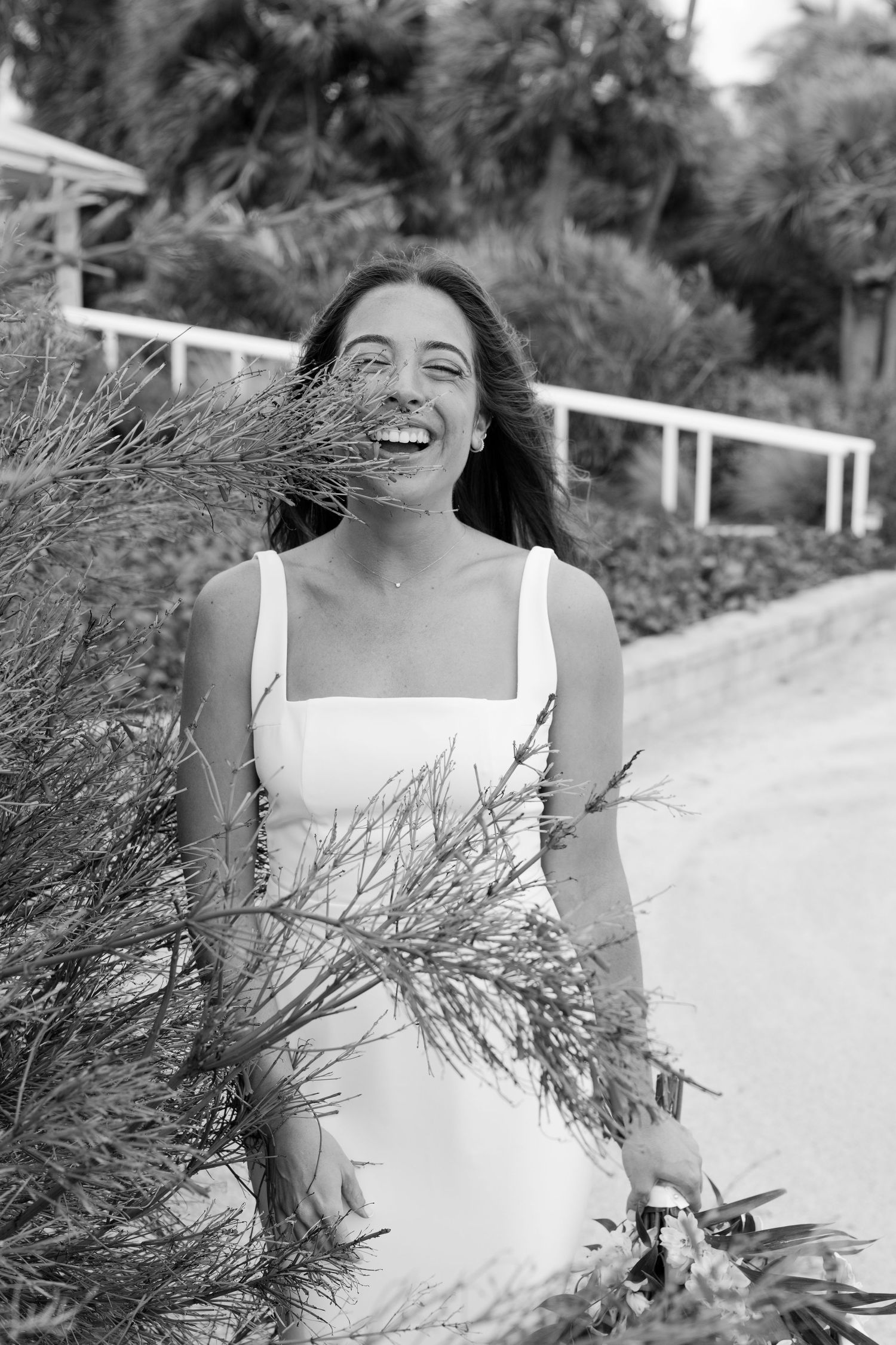 Black and white portrait of a laughing bride in a white dress standing near lush greenery outdoors.