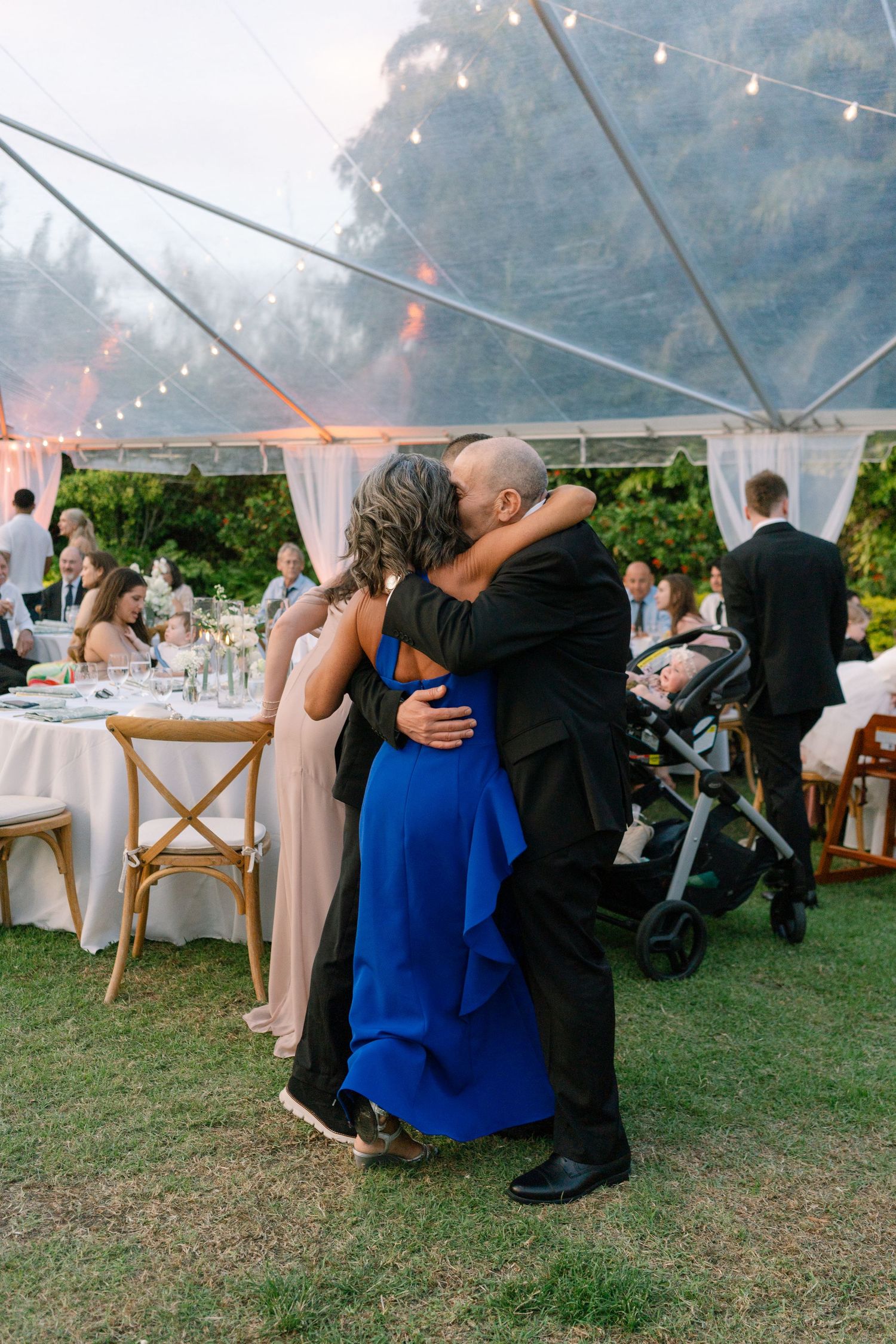 A couple shares a warm embrace on the grass under a clear tent at an elegant outdoor wedding reception.