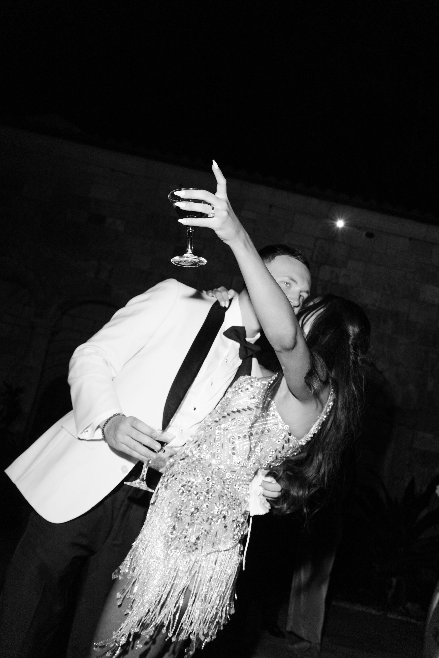 Elegant couple dancing at night, woman in sequined fringe dress holding champagne glass, man in white tuxedo jacket, black and white photo.