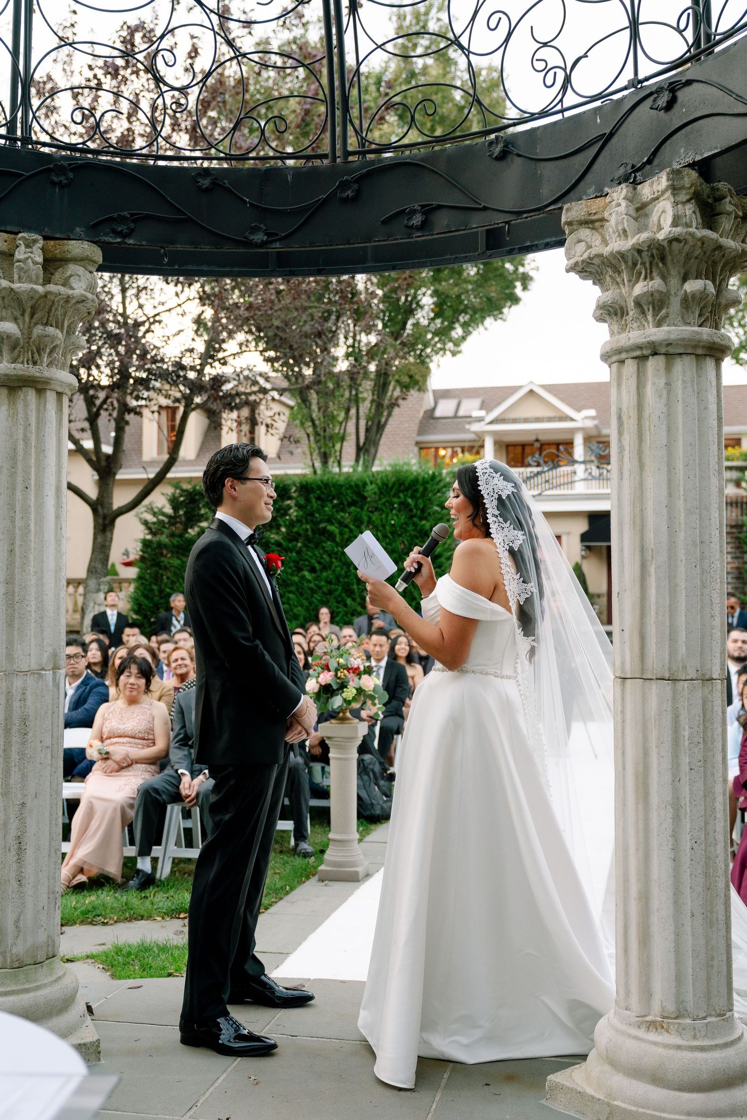 A wedding ceremony takes place under an ornate gazebo with classical columns.