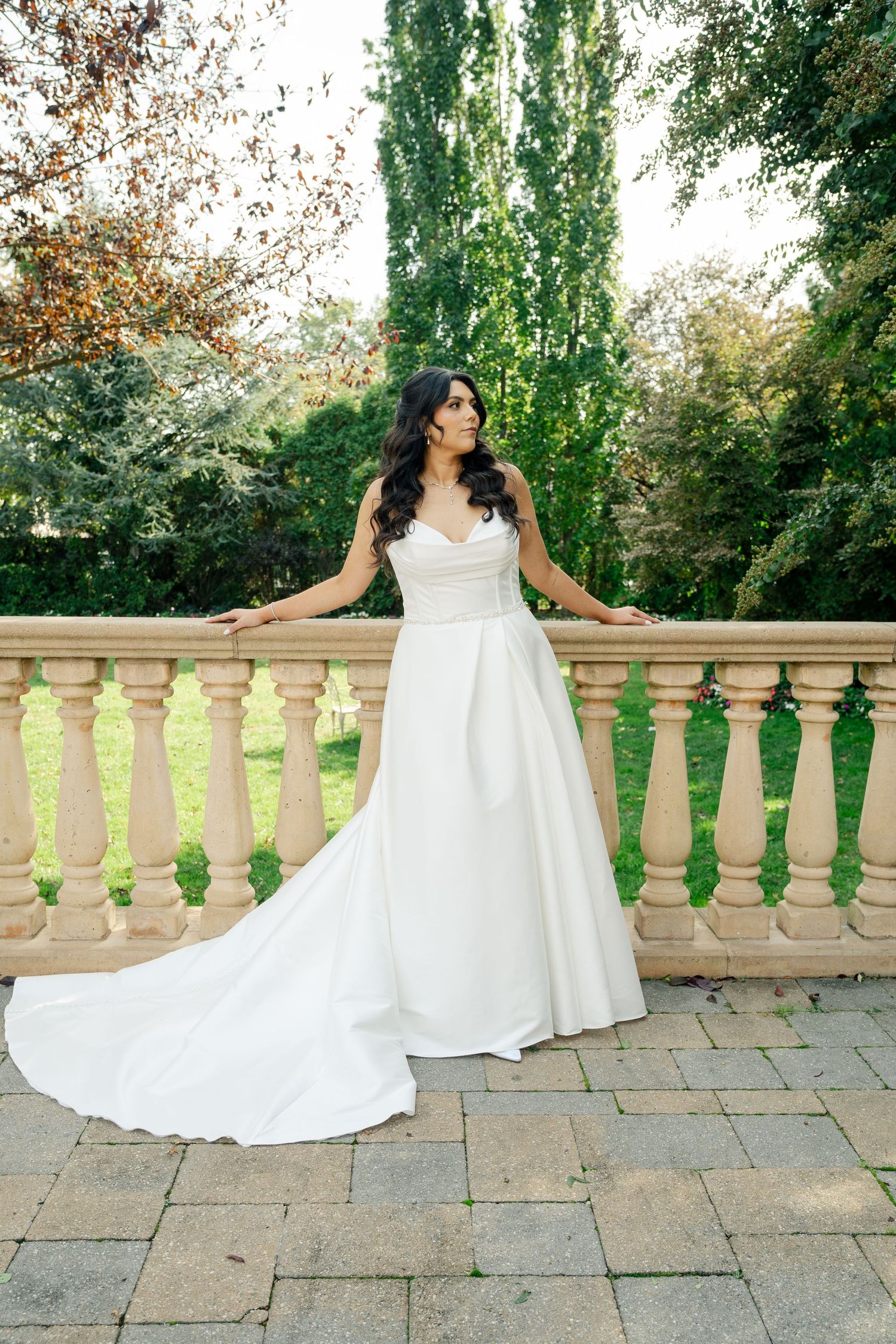 Bride in flowing white dress poses along stone balustrade in garden setting.
