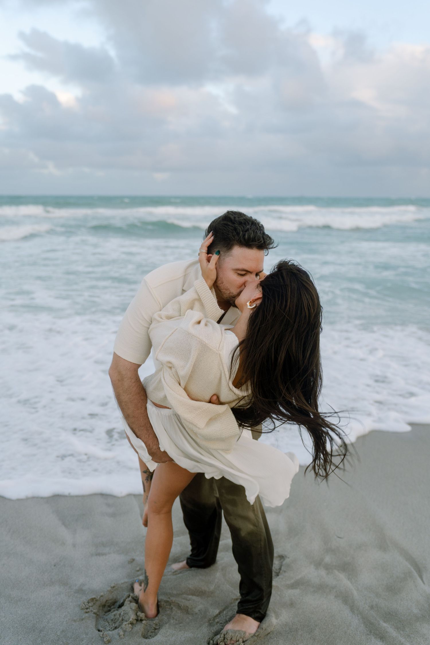 A romantic couple shares an intimate moment on a cloudy beach with crashing waves in the background.
