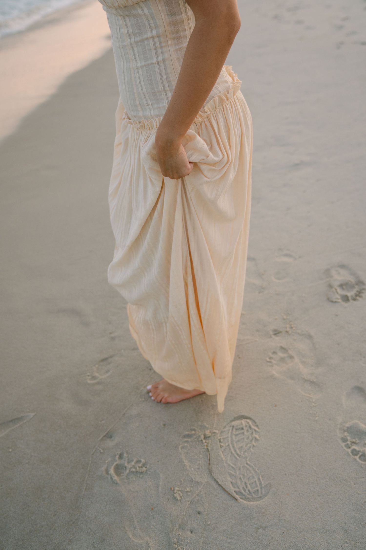 Bare feet walking on sandy beach leaving footprints in cream-colored flowing dress, captured in soft light.