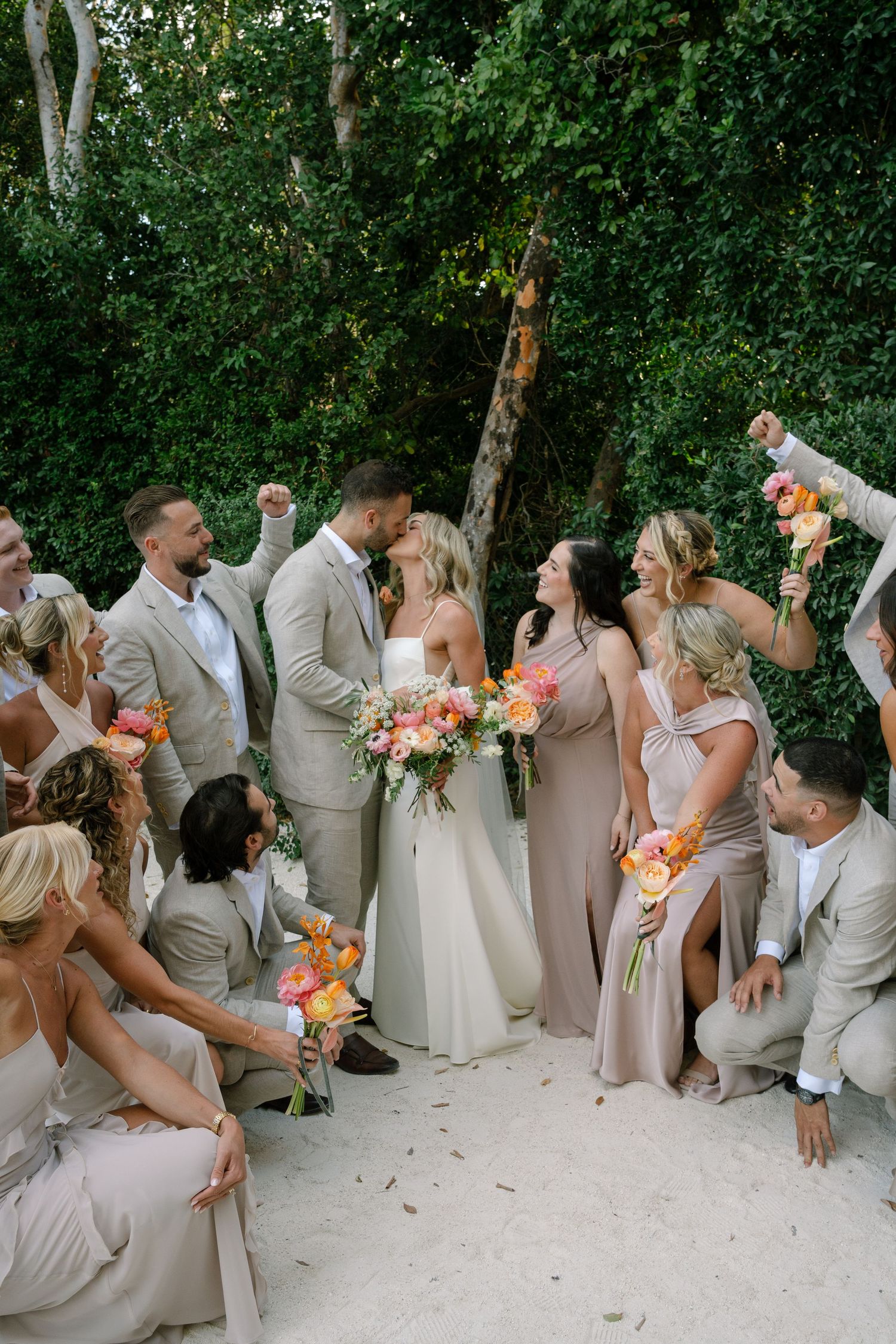 Wedding party in light colored attire poses playfully in a line against lush green foliage backdrop.
