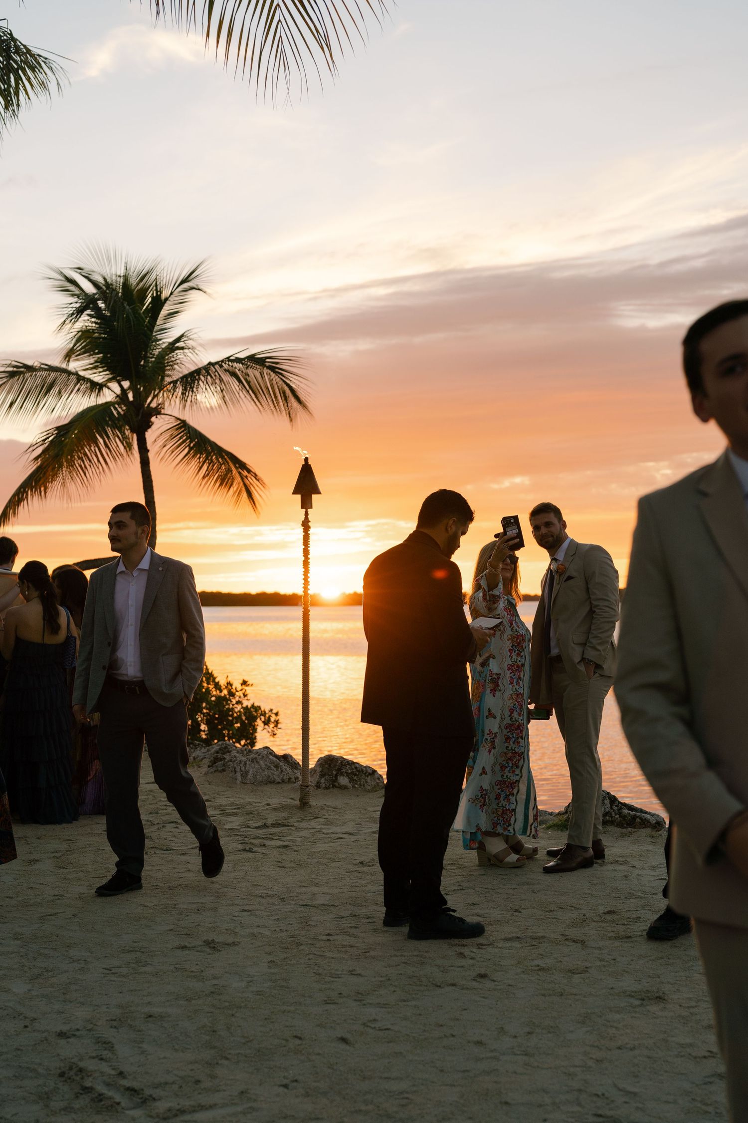 Silhouettes of wedding guests on a beach at sunset with palm trees and orange sky.