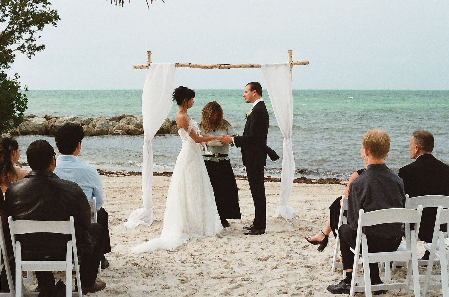 Couple exchanging vows at a romantic beach wedding ceremony under a rustic wooden arch with white fabric.