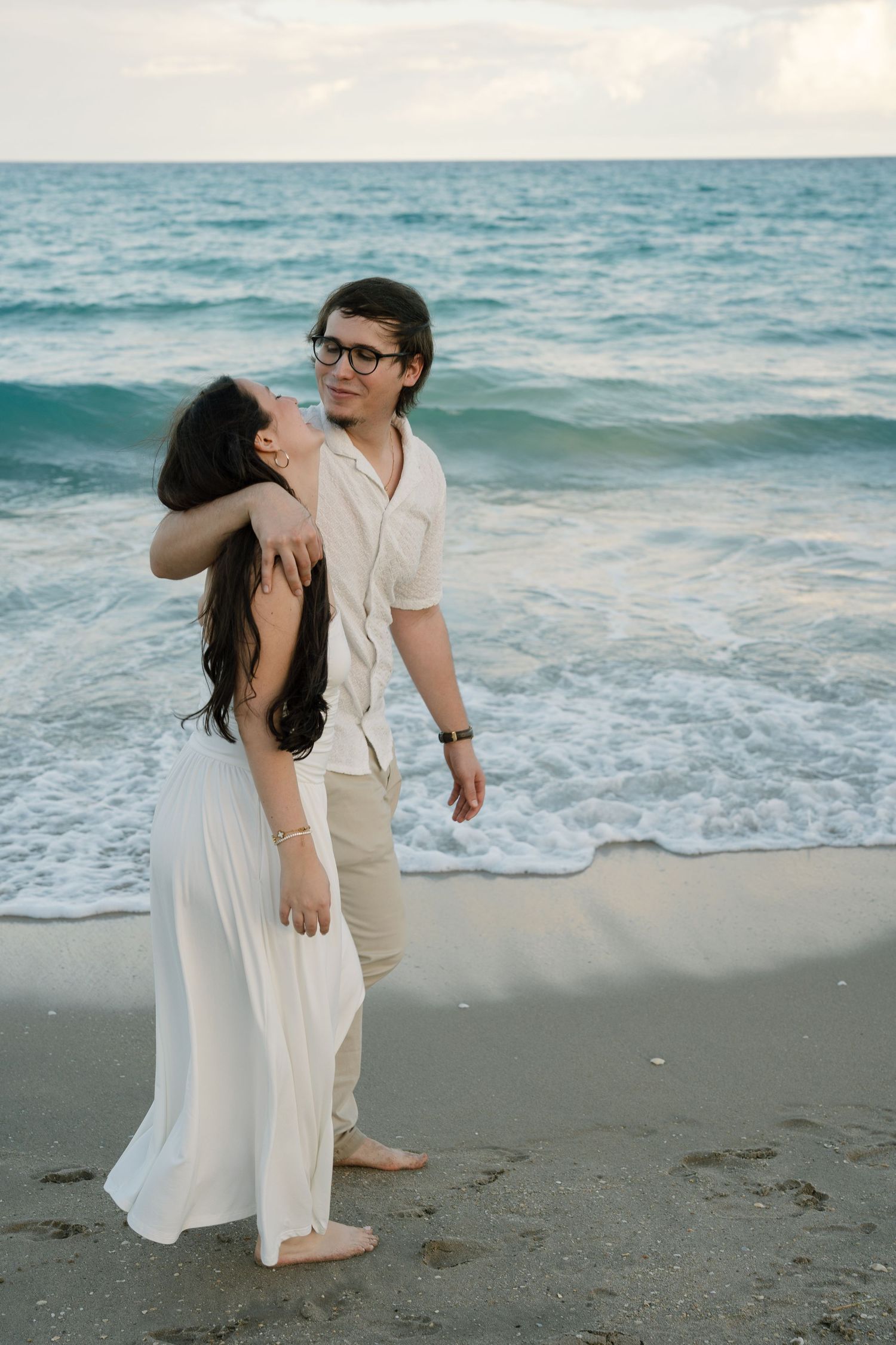 A couple in white and beige outfits share romantic moments on a beach with waves in the background at sunset.