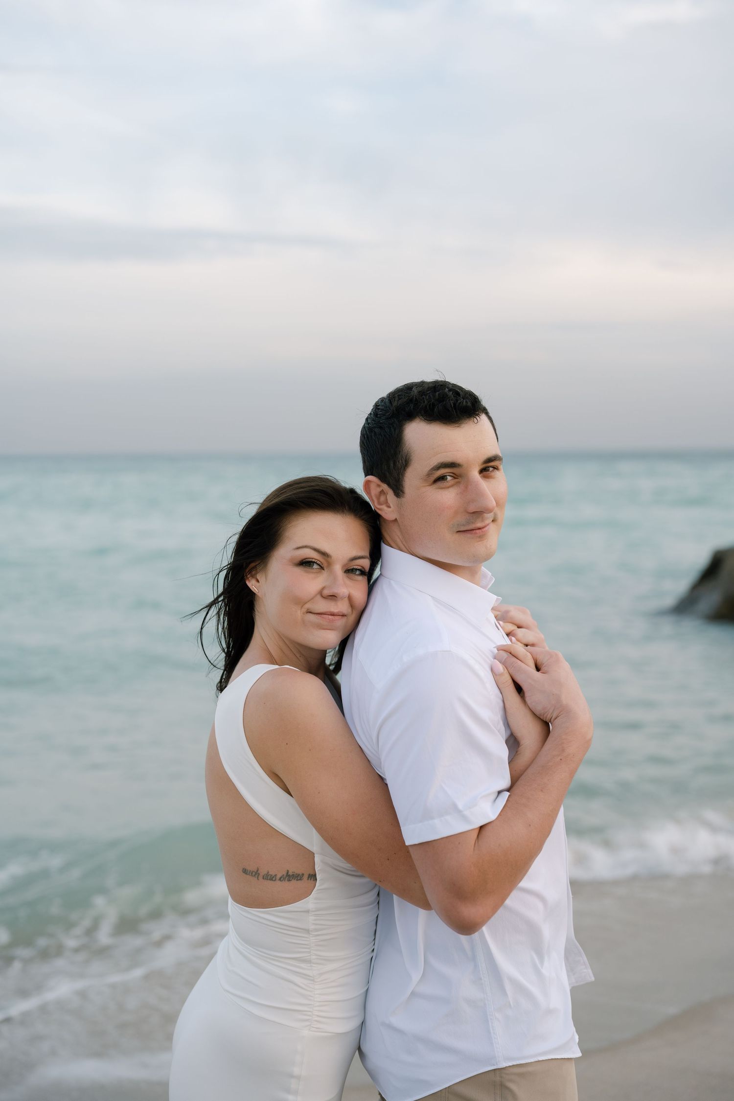 A couple in white clothing embraces tenderly on a peaceful beach with the ocean horizon behind them.