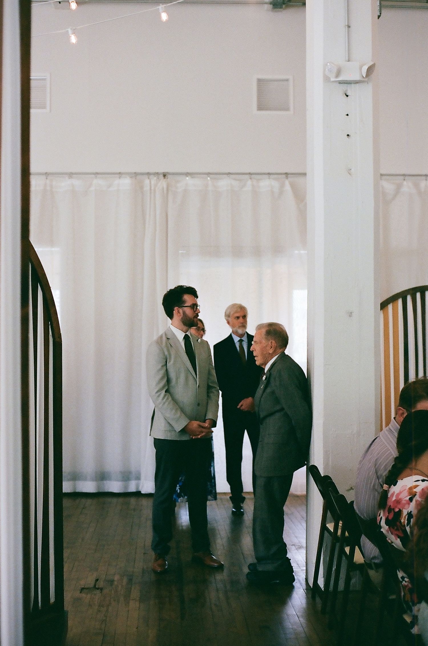 A minimalist indoor venue with white curtains and wooden floors where guests gather in formal attire.