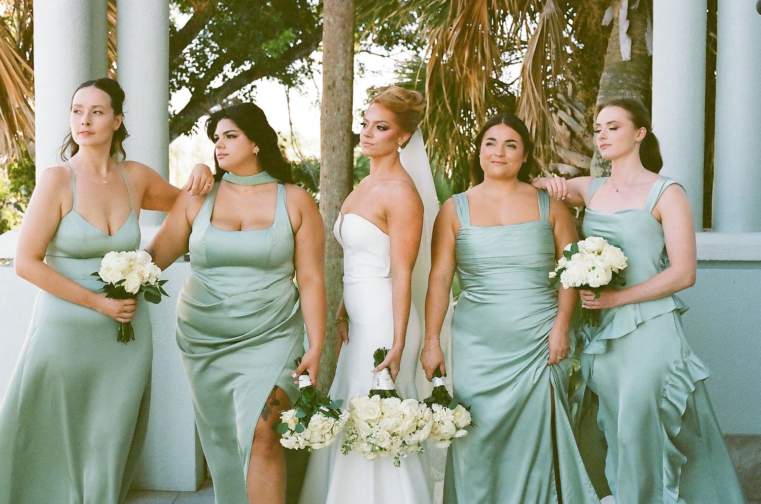 Wedding party in seafoam green dresses pose elegantly on a porch with white columns and tropical foliage.