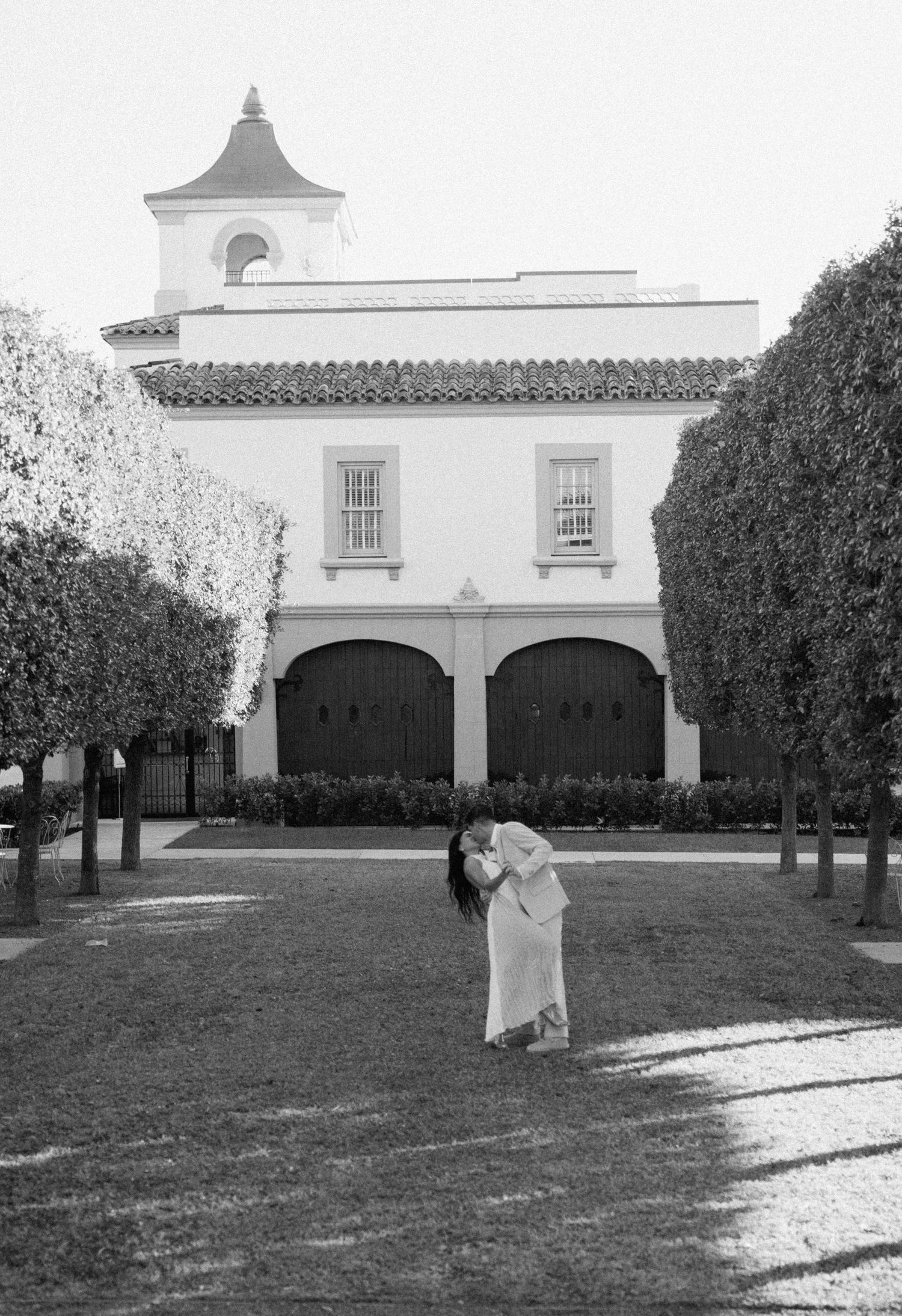A black and white romantic moment captured in front of a Spanish colonial style building with arched doorways and a bell tower.