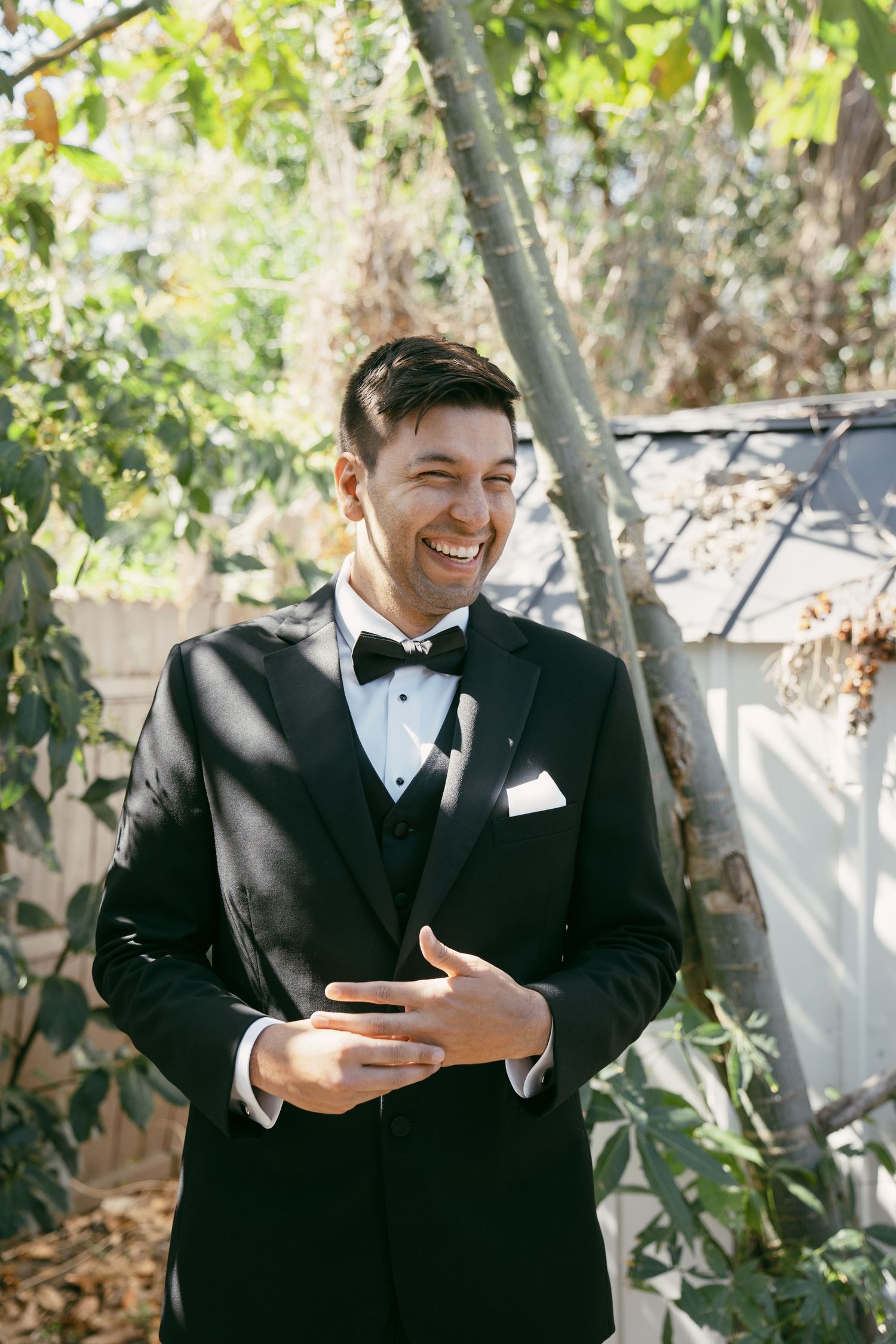 A joyful groom in a classic black tuxedo and bow tie laughs while standing in a garden setting on his wedding day.