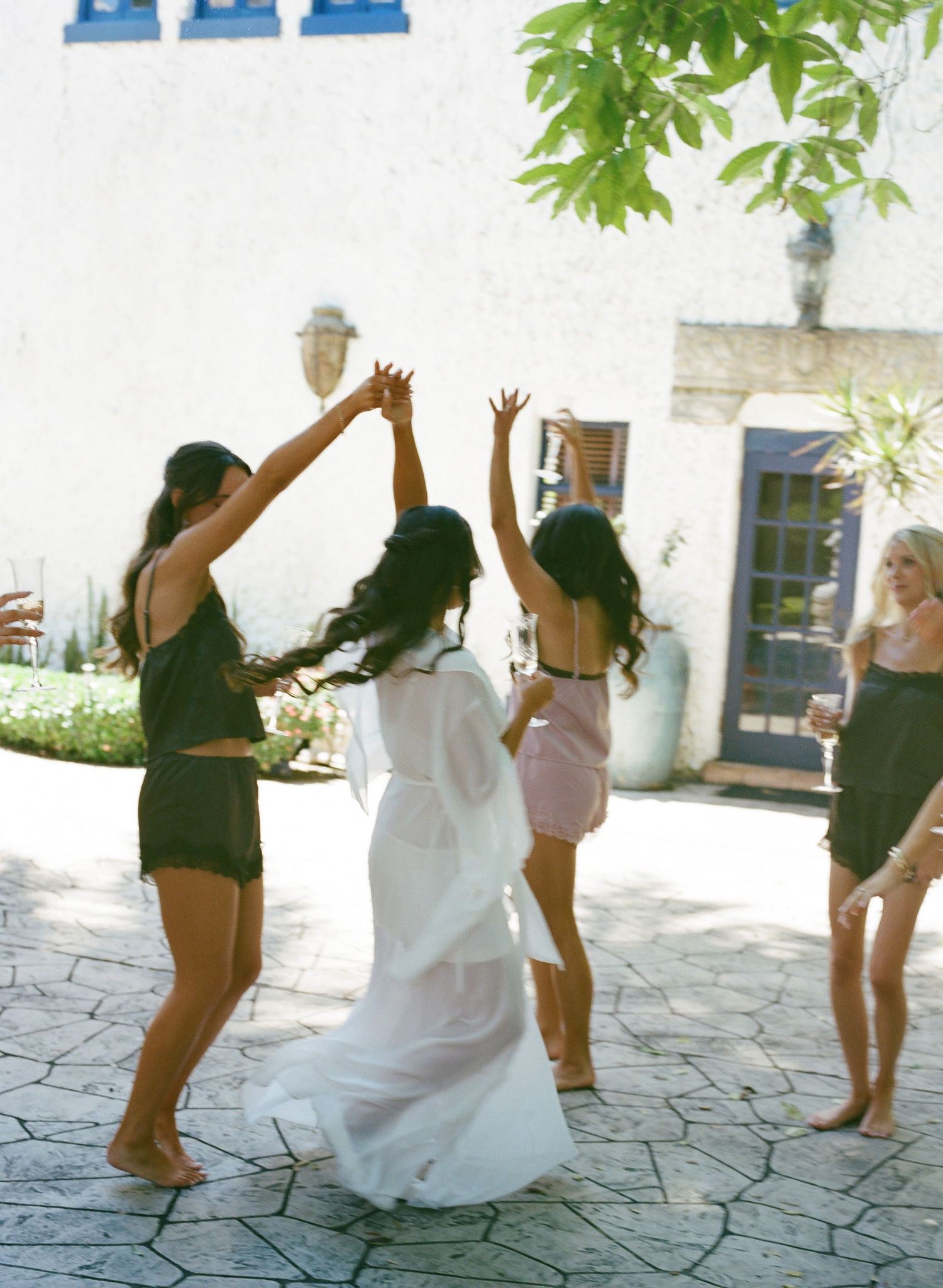 Bride in flowing white gown dances outdoors with bridesmaids in dark outfits at sunny wedding celebration.