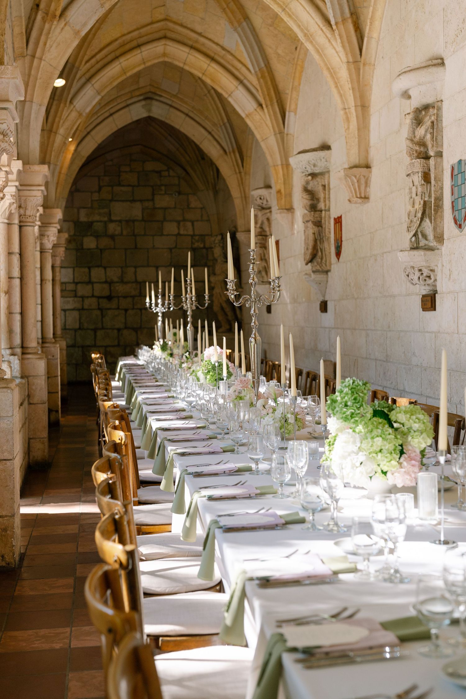Elegant wedding reception in a Gothic stone cloister with long table, candelabras, white florals, and green napkins.