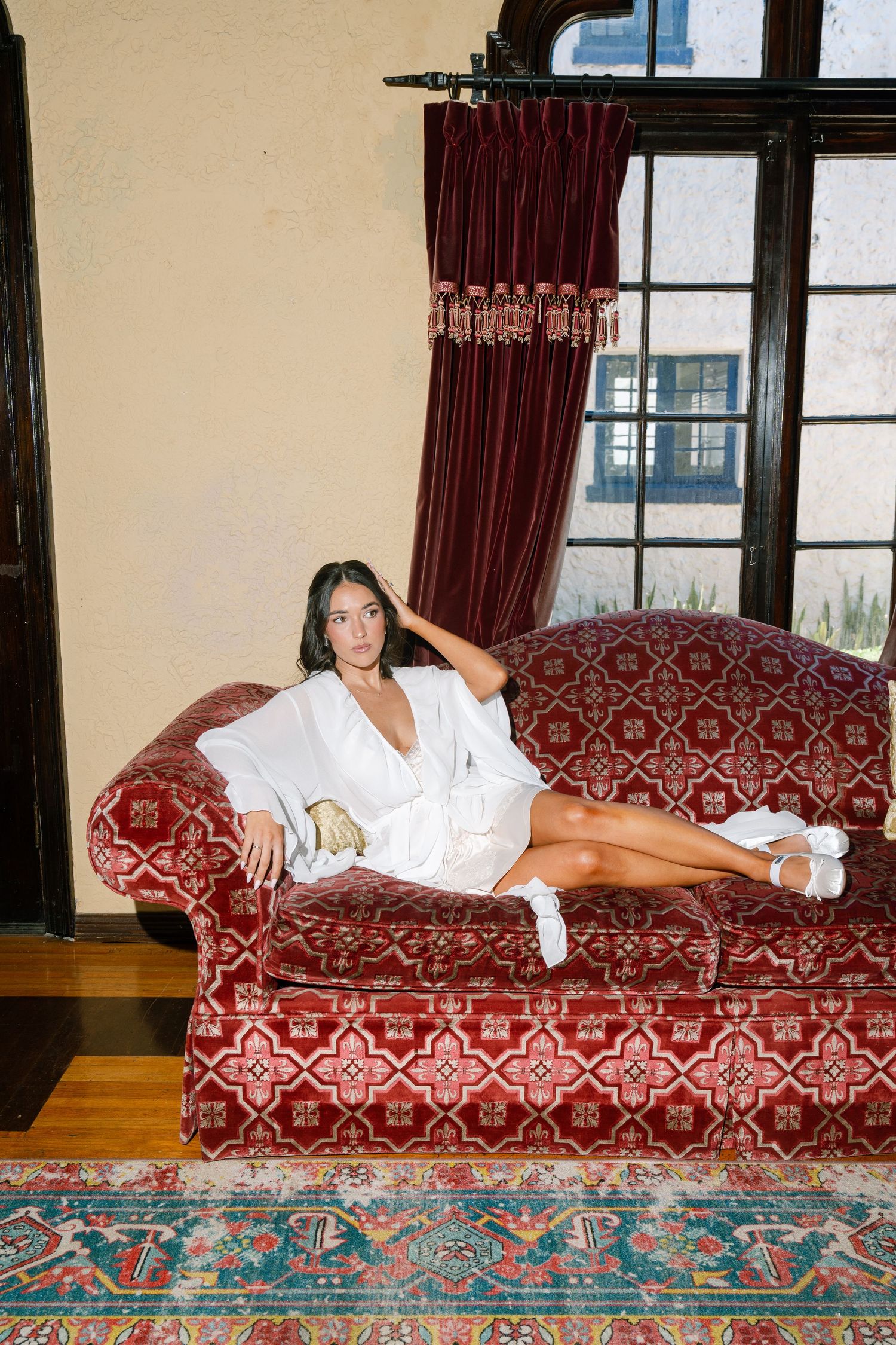 Bride in white robe lounging on ornate red velvet sofa in elegant room with dark curtains and hardwood floors.
