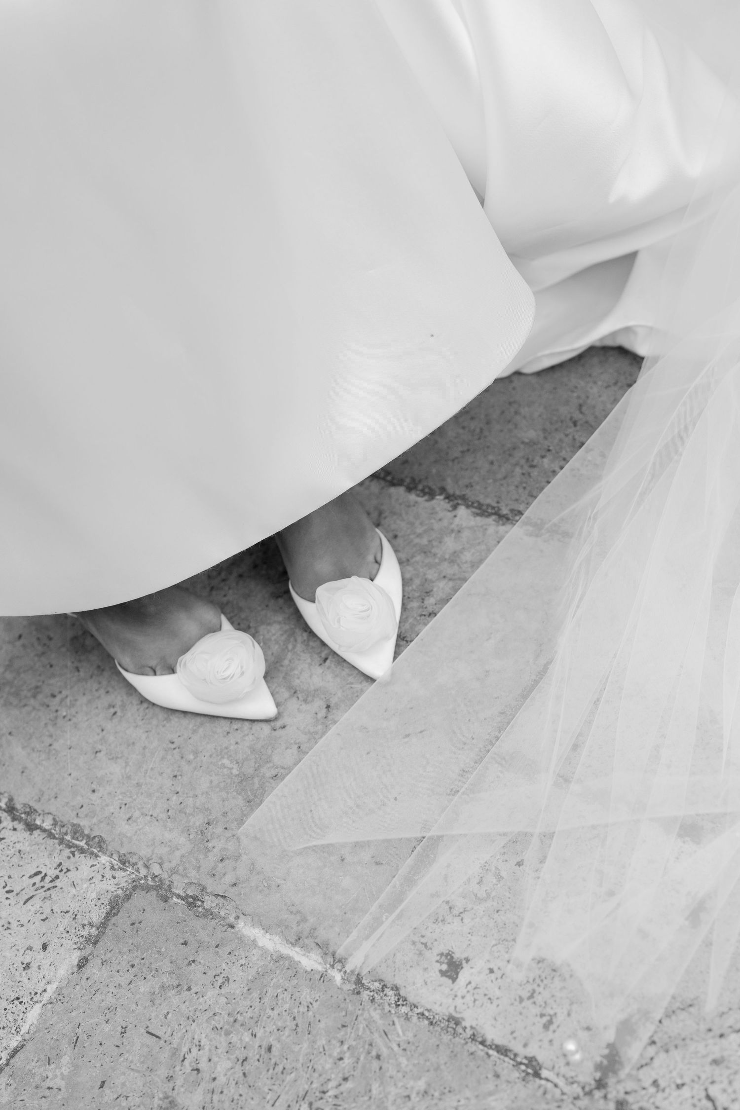 Black and white photo of a bride wearing white heels with floral detail, wedding dress hem and veil visible on stone floor.