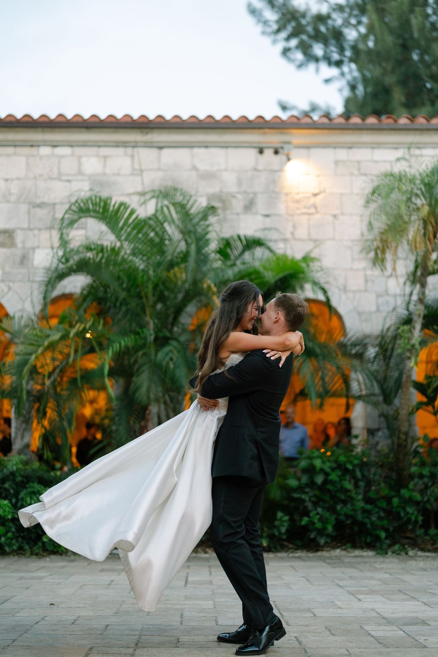 Groom lifts bride in white gown outside a Mediterranean-style venue surrounded by tropical palm trees at dusk.