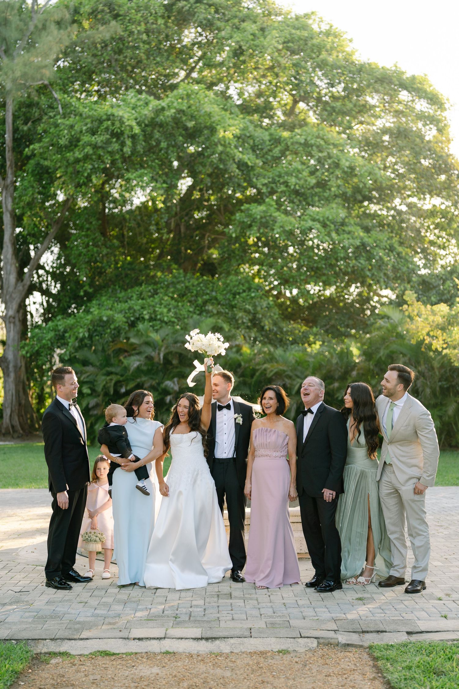 Wedding party poses outdoors with bride raising bouquet beneath a large green tree.