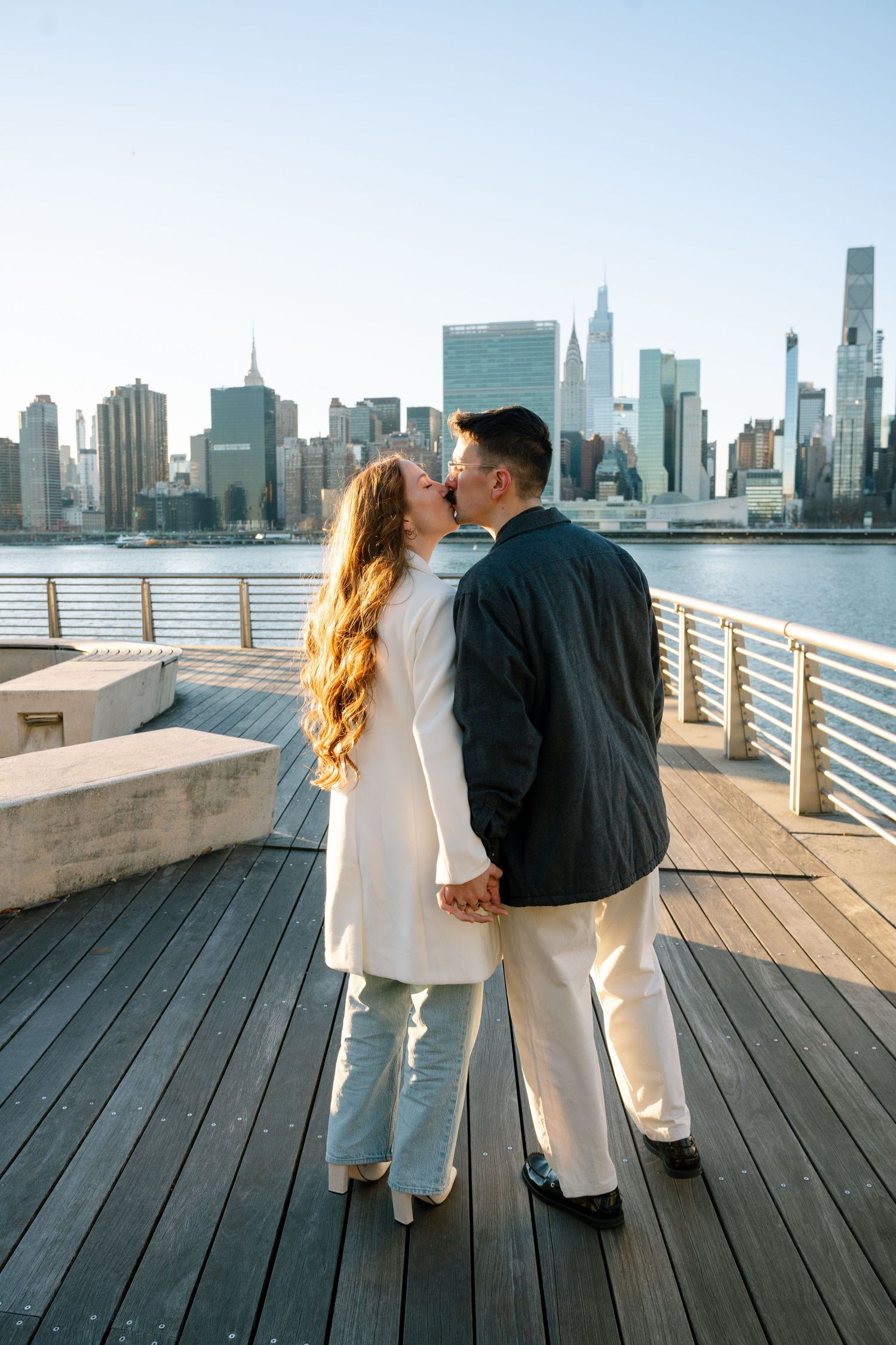 Two people share a romantic moment on a wooden pier with the Manhattan skyline as their backdrop.