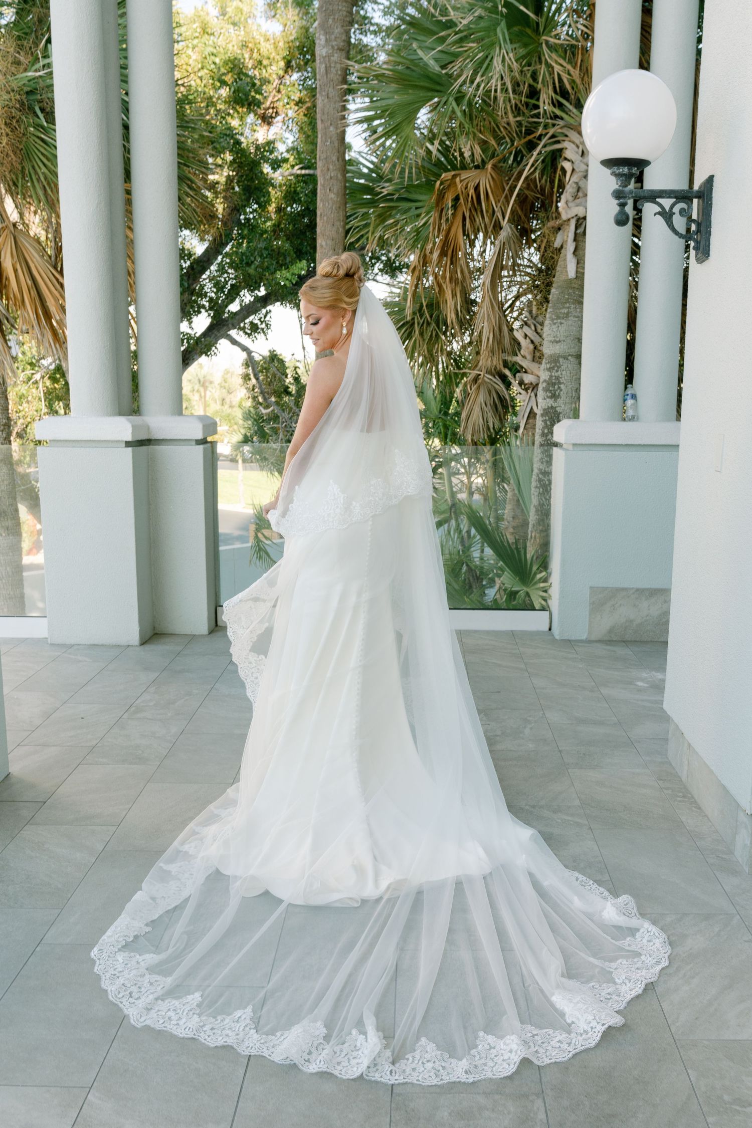 White wedding gown with lace trim and flowing train photographed on a columned porch.