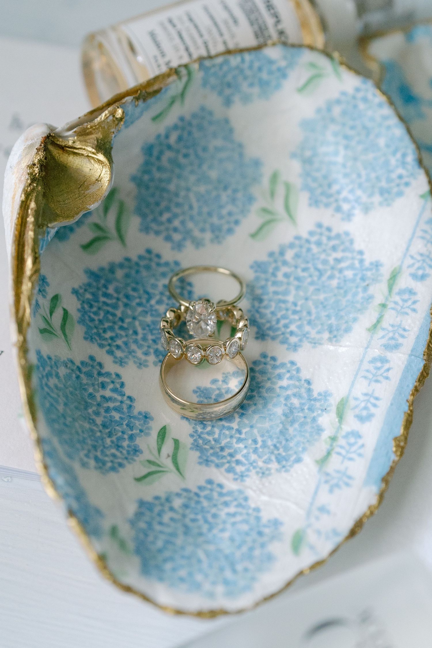 Gold rings displayed in a decorative oyster shell dish with blue hydrangea floral pattern and gold leaf trim.
