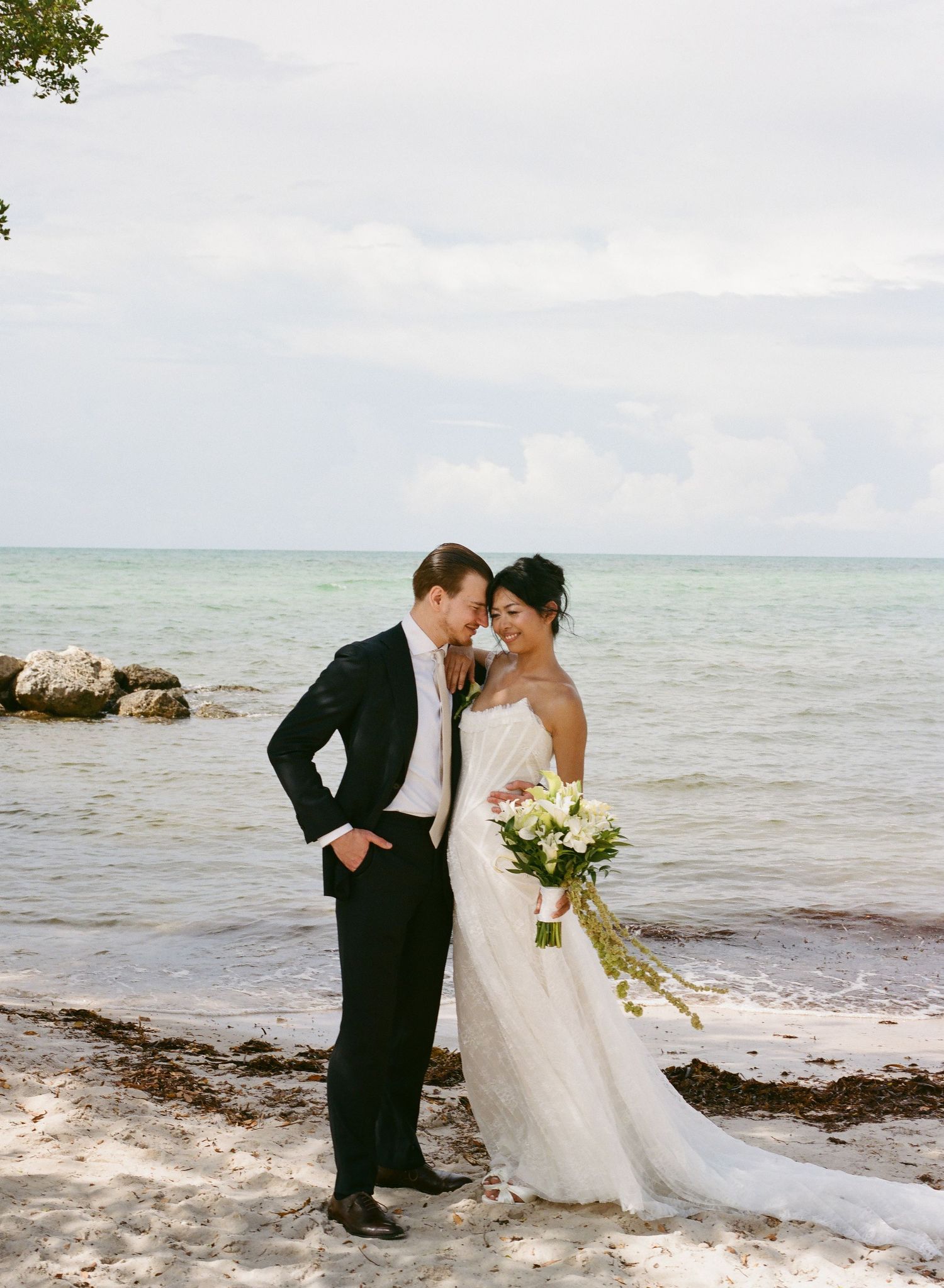 Bride and groom share a romantic moment on a sandy beach, with calm ocean waters and cloudy skies behind them.