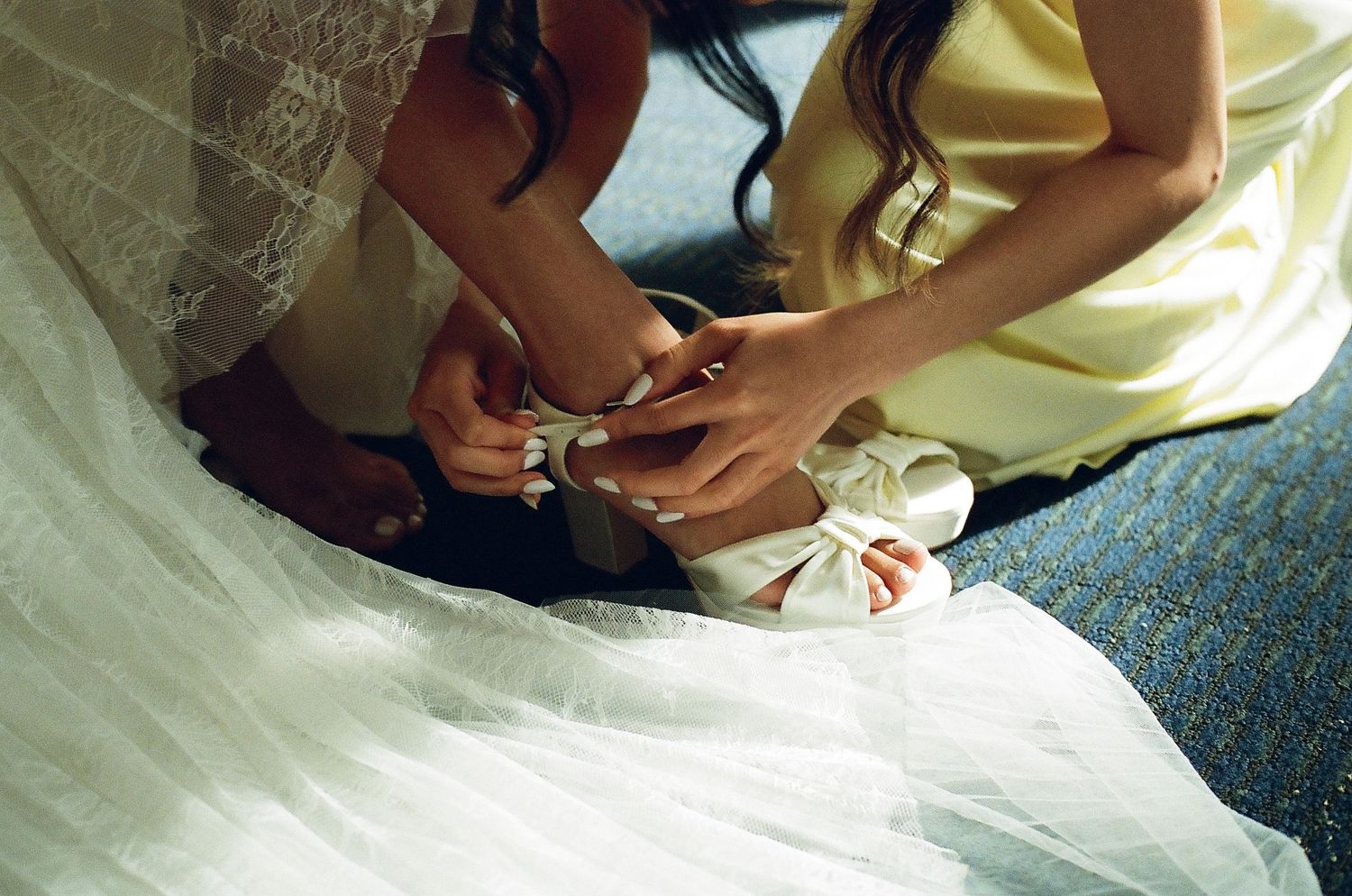 Bridesmaid in yellow dress helping bride fasten white wedding shoes before the ceremony.