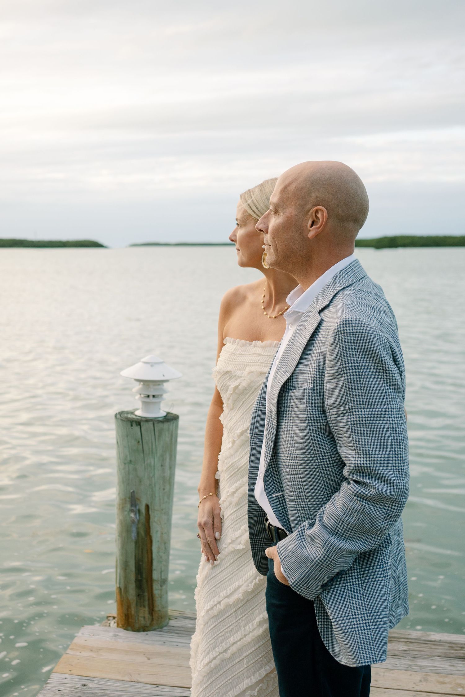 A couple stands on a wooden dock overlooking calm waters at sunset, with a nautical post light in the foreground.