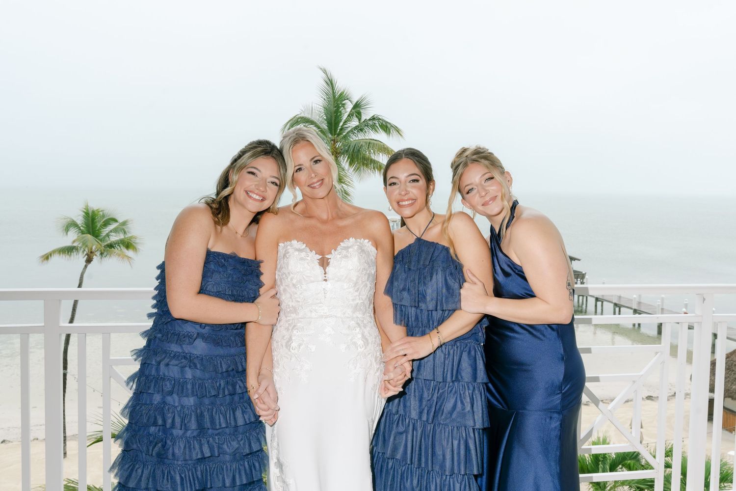 Bridal party in navy blue dresses poses together on a sunny beach with white picket fence.