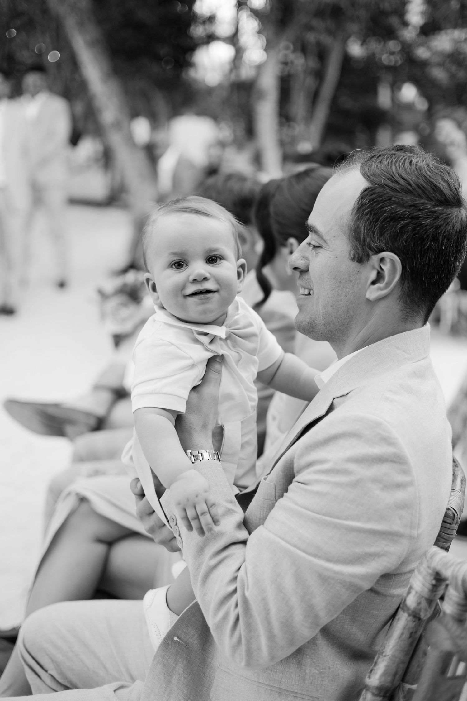 A black and white photo of a guest in a light suit holding a small child at an outdoor event.
